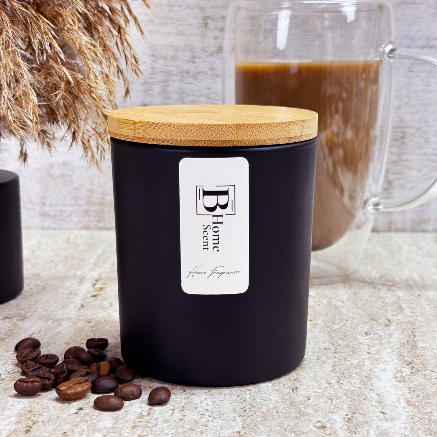 A black Coffee Cake scented candle with a wooden lid, labelled 'B Home Scent,' displayed on a marble surface with coffee beans scattered around and a glass of coffee in the background.