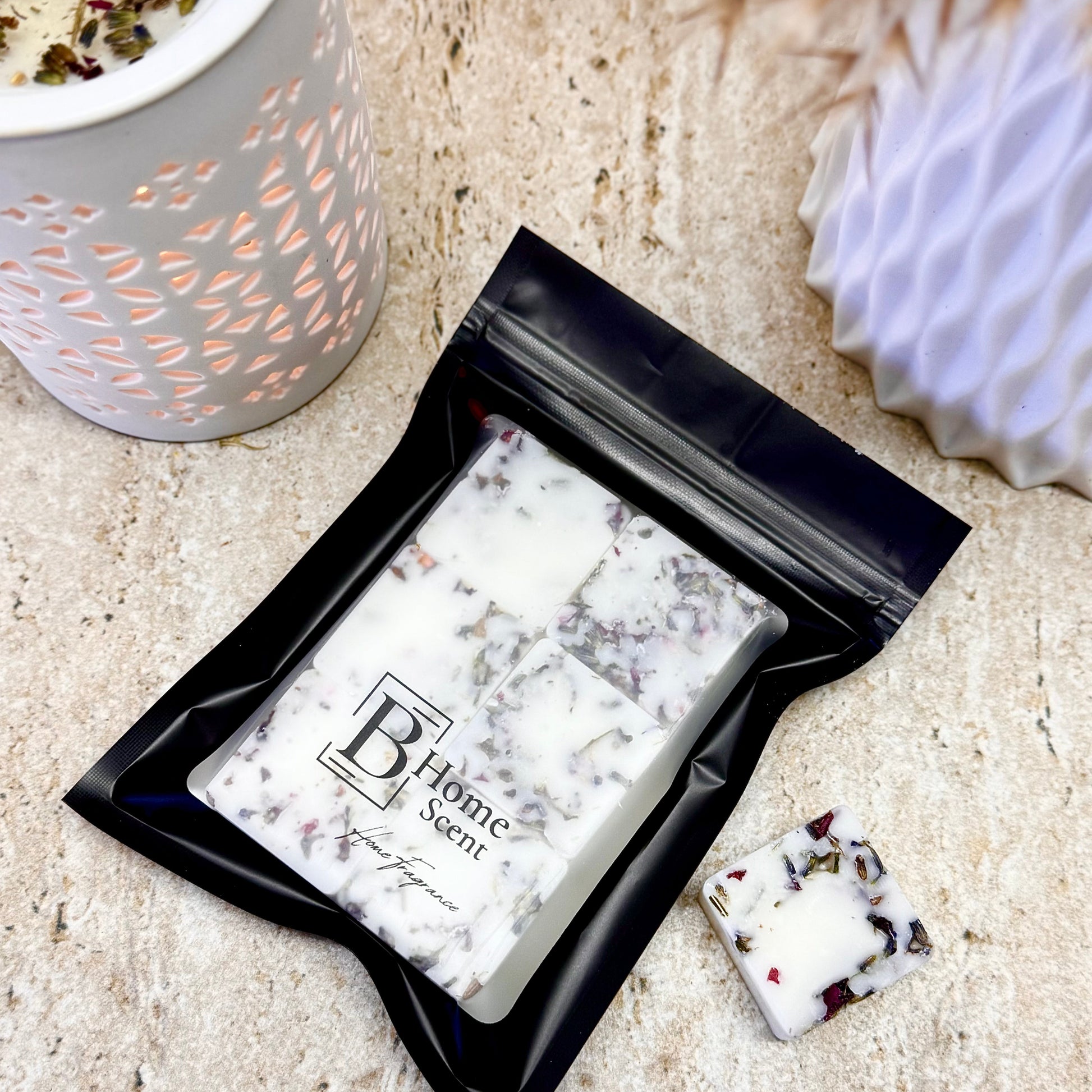 Coconut Milk & Patchouli Botanical Wax Melt Bar with Dried Petals in Black Resealable Pouch, Shown Beside Ceramic Wax Burner on Stone Surface