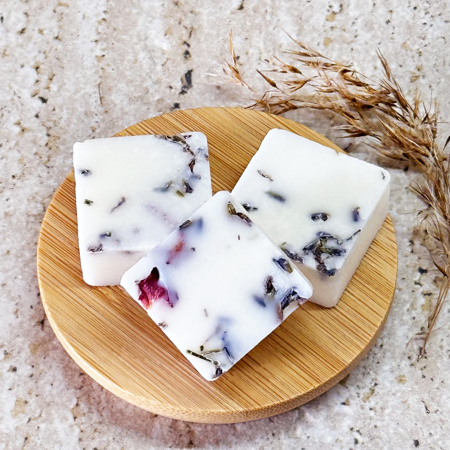 Three blackberry and cedarwood wax melts with dried lavender and rose petals are displayed on a round wooden plate placed on a light marble surface. Dry grass stems lie beside the plate, adding a natural touch to the setting.