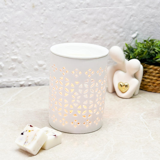 A white ceramic burner with an intricate geometric cut-out design, glowing softly with a warm light. The burner is styled with wax melts in the foreground and decorative elements, including a green plant and a white figurine, in the background.