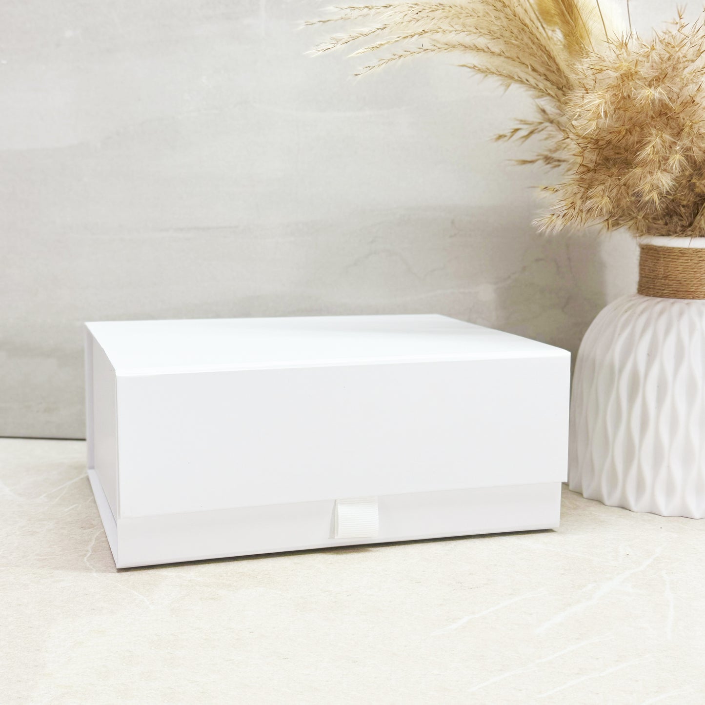 A sleek, white laminated magnetic gift box placed on a neutral-coloured surface, with a vase of dried flowers in the background.