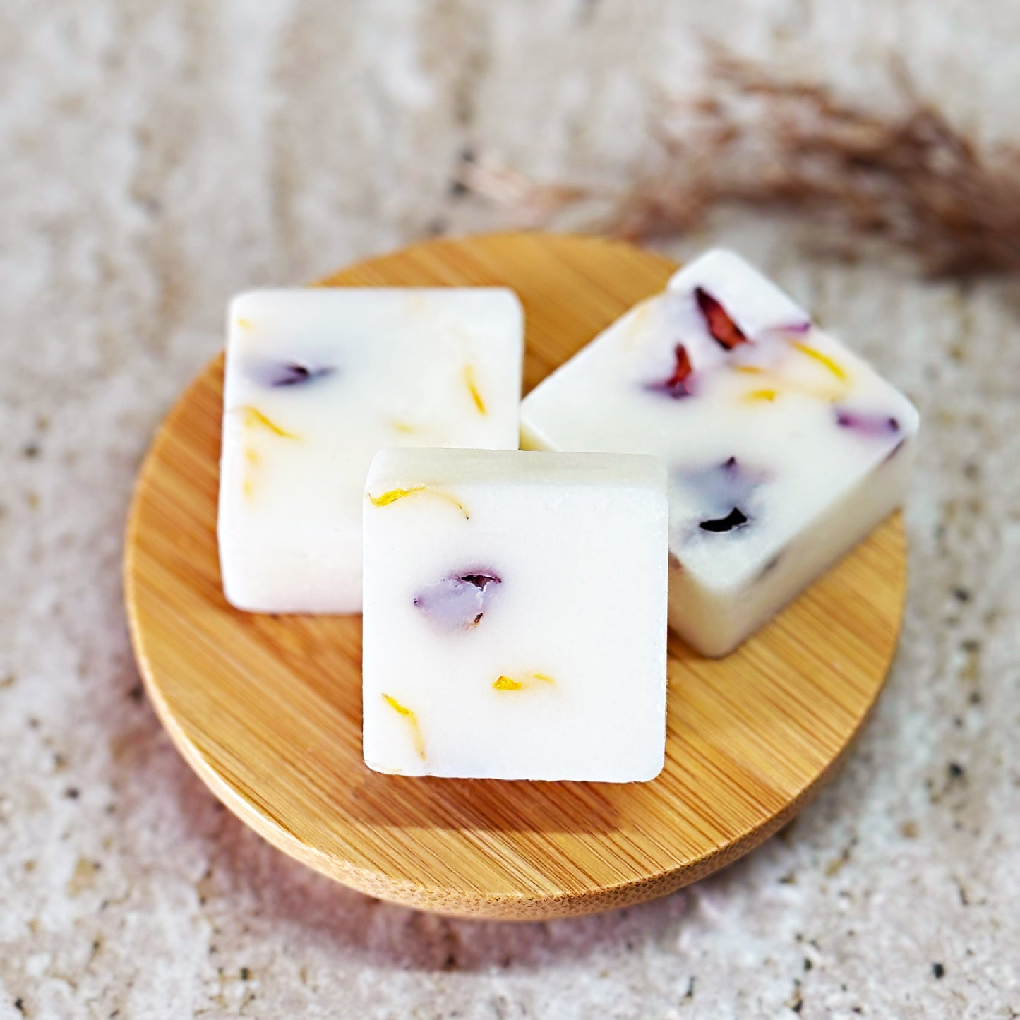 Three Pumpkin Spice Latte wax melts with dried calendula and rose petals are displayed on a round wooden plate placed on a light marble surface. Dry grass stems lie beside the plate, adding a natural touch to the setting.