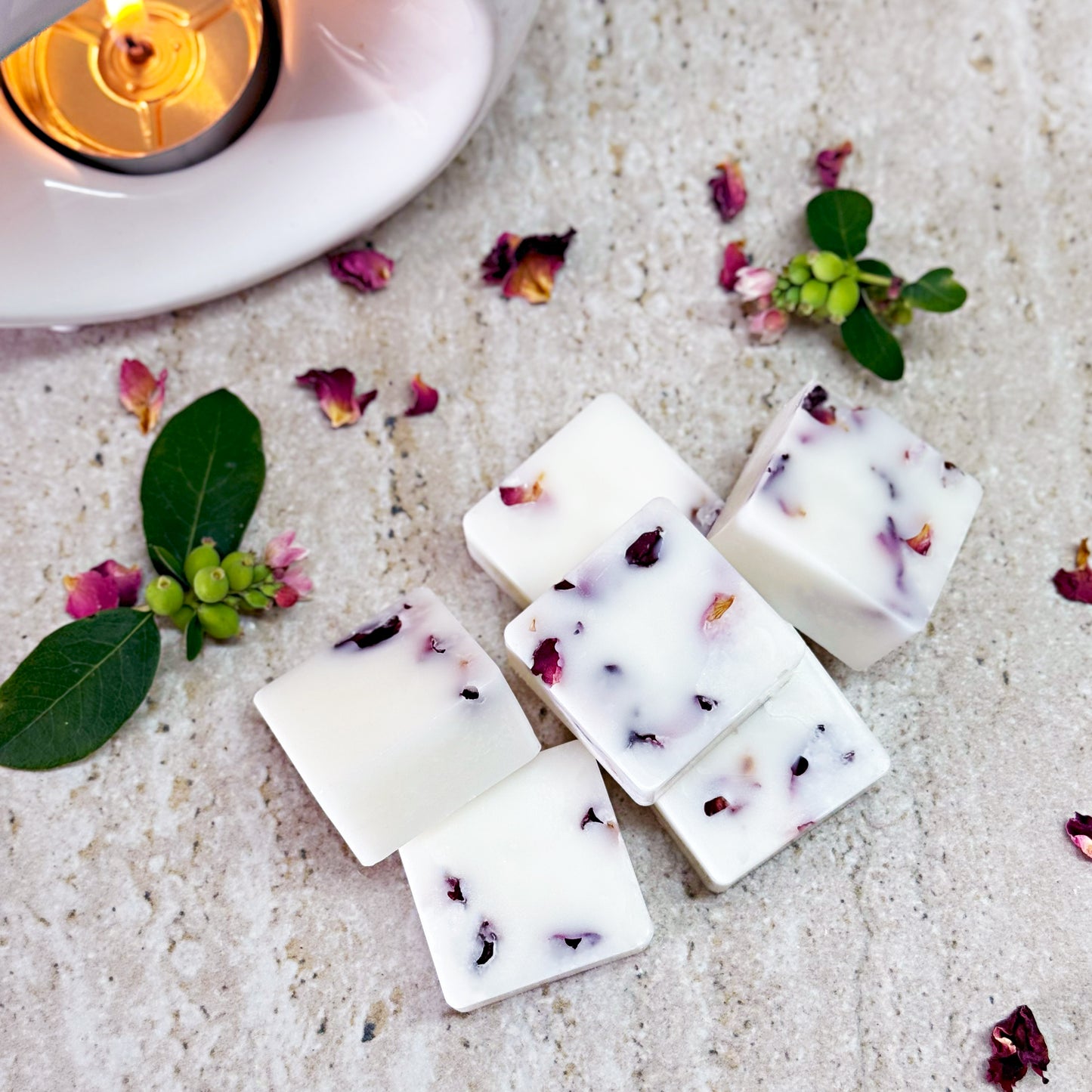 Six apple blossom prosecco botanical wax melts with dried rose petals arranged on a light textured surface, surrounded by small green leaves, pink flowers, and a lit tea light in a white wax melt burner.
