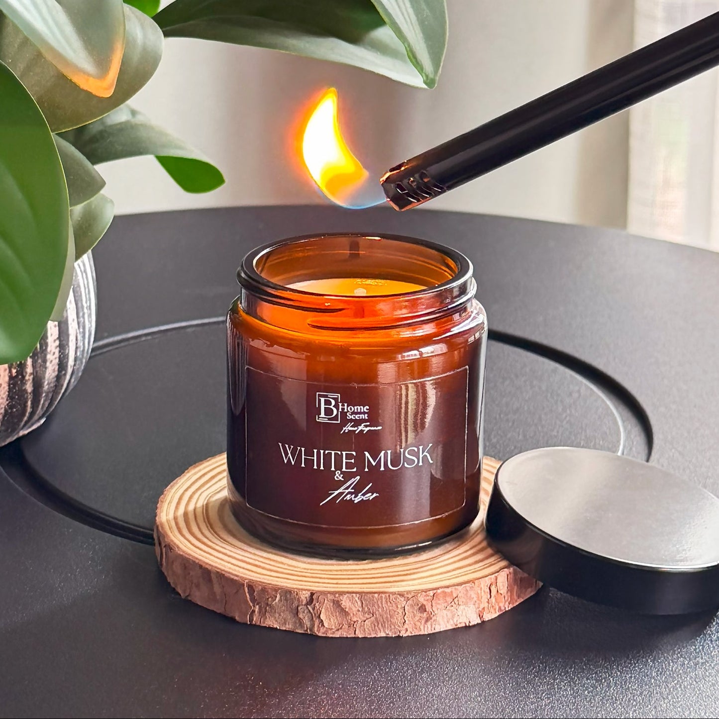 White Musk and Amber Soy Candle being lit with a match on a dark surface next to a plant.
