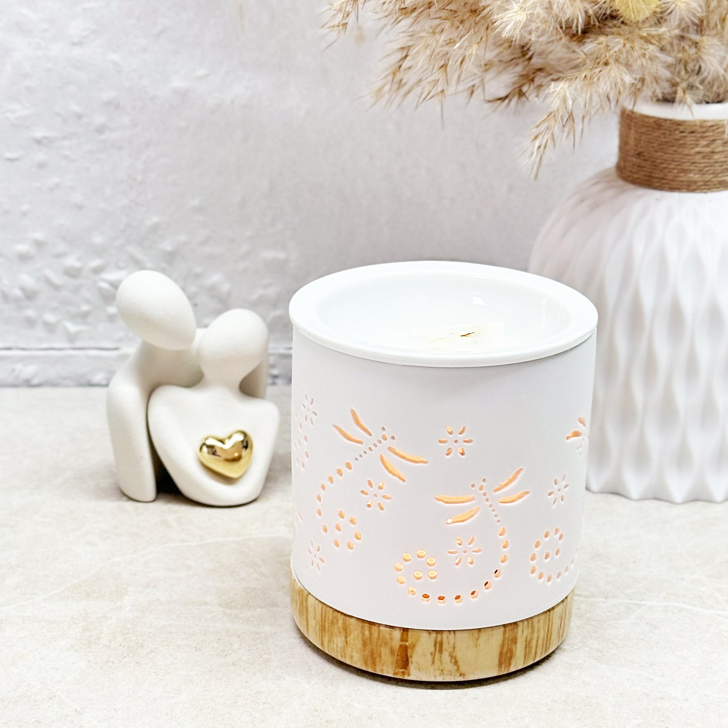 A white ceramic burner with delicate dragonfly and floral cut-out patterns, resting on a ceramic base with a wood-effect finish. The burner is lit, emitting a warm glow, and is styled with a minimalistic white vase and a figurine in the background.