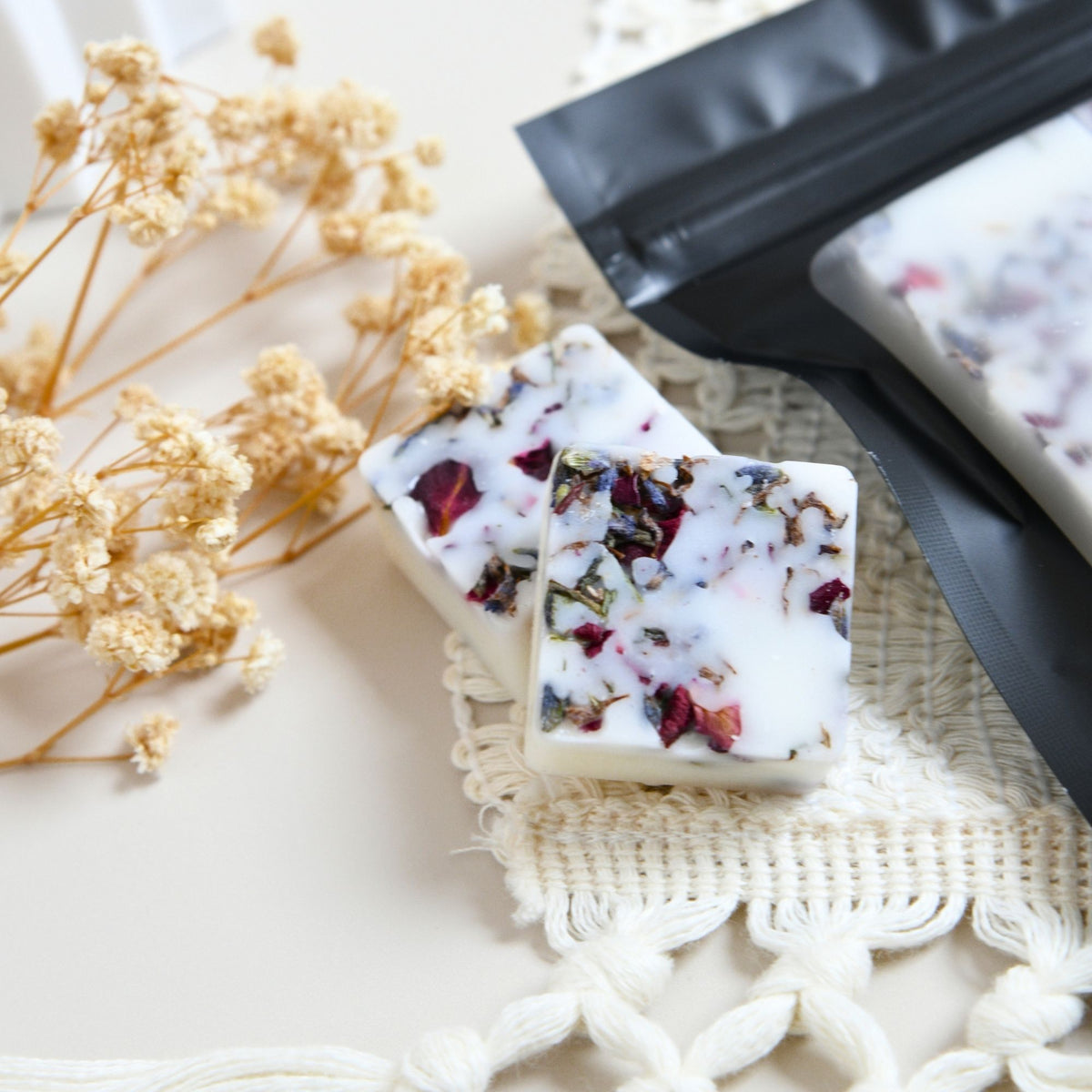 Two square cubes of handmade wax melts with dried flower petals and lavender embedded in a white wax melts, displayed next to a black resealable pouch on a cream lace runner with dried flowers in the background.
