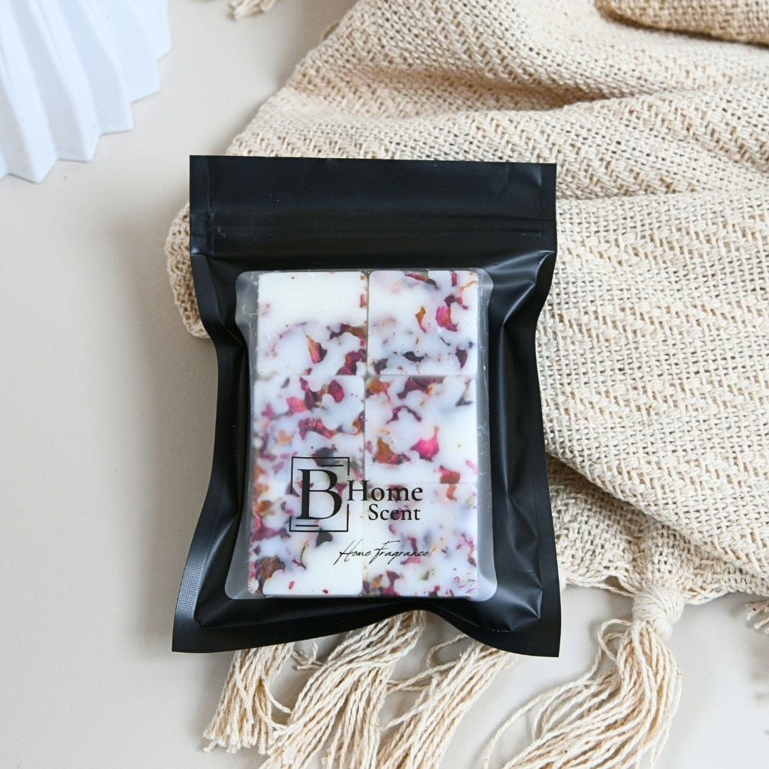A black resealable pouch containing a white wax melt  bar with red rose petals, placed on a cream surface with a beige knitted blanket and tassels behind it. The pouch label reads 'B Home Scents'.