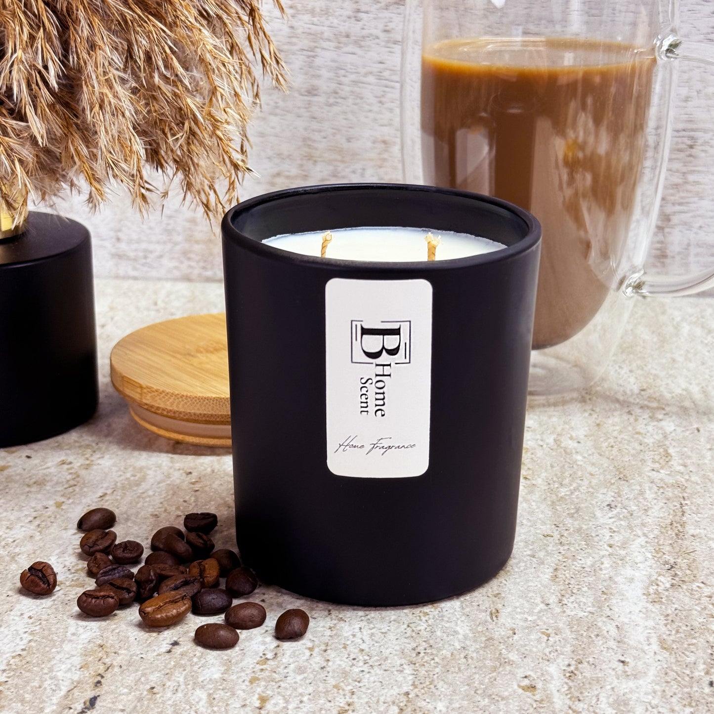 A black Coffee Cake scented candle with two wicks, labelled 'B Home Scent,' placed on a marble surface alongside coffee beans and a glass of coffee in the background.