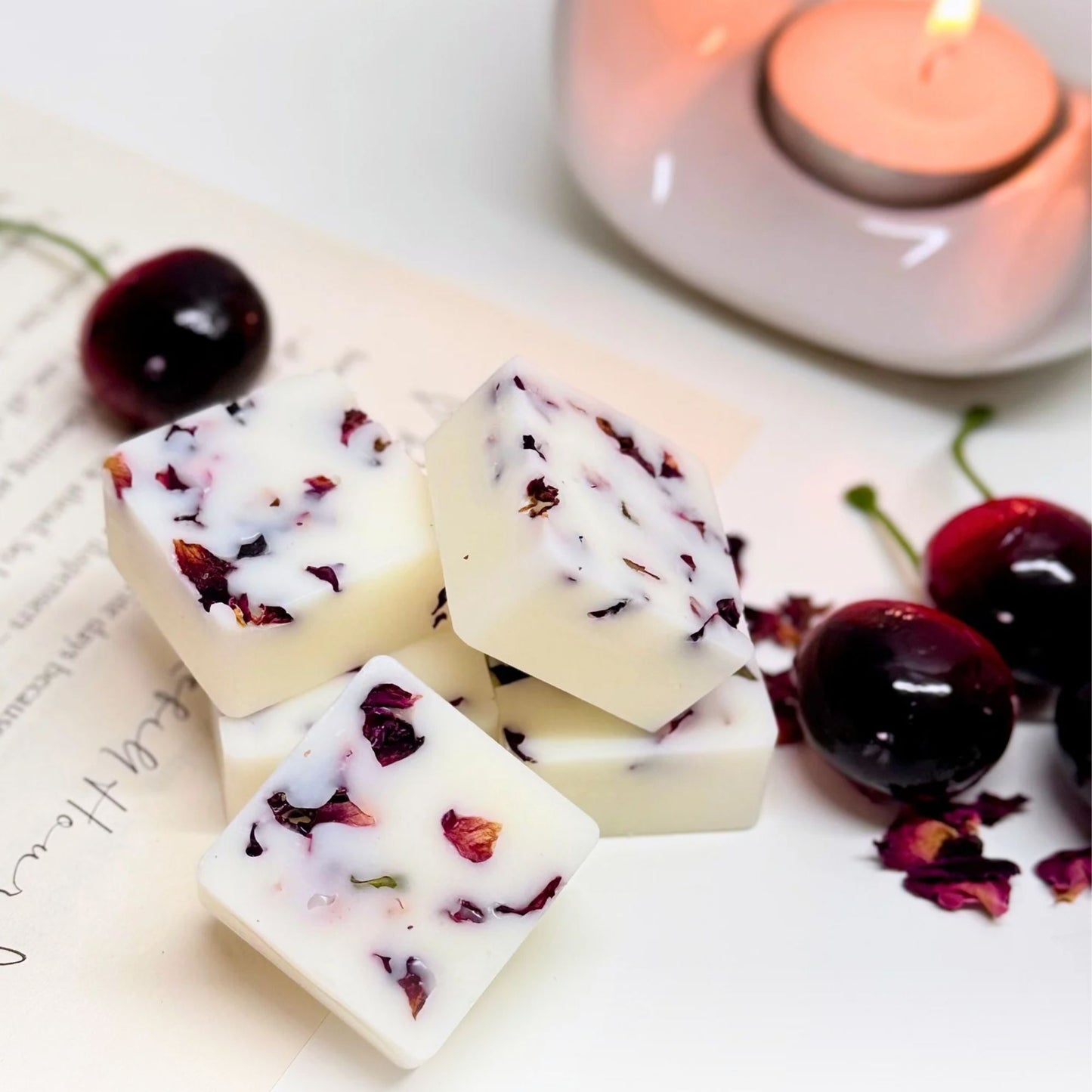 Close-up of Black Cherry botanical wax melts with dried rose petals embedded in creamy white wax. The melts are stacked on a white surface with scattered rose petals and fresh black cherries nearby. A lit tealight candle in a white ceramic wax burner adds a warm, cosy glow in the background.