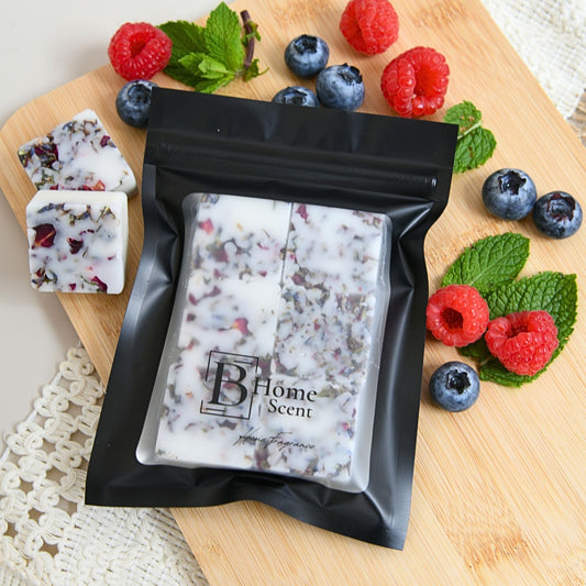 Blackberry and cedarwood wax melts in  black resealable pouch labeled 'B Home Scent' containing a white wax melt with dried floral pieces, styled on a wooden board with raspberries, blueberries, mint, and two loose cubes beside it.