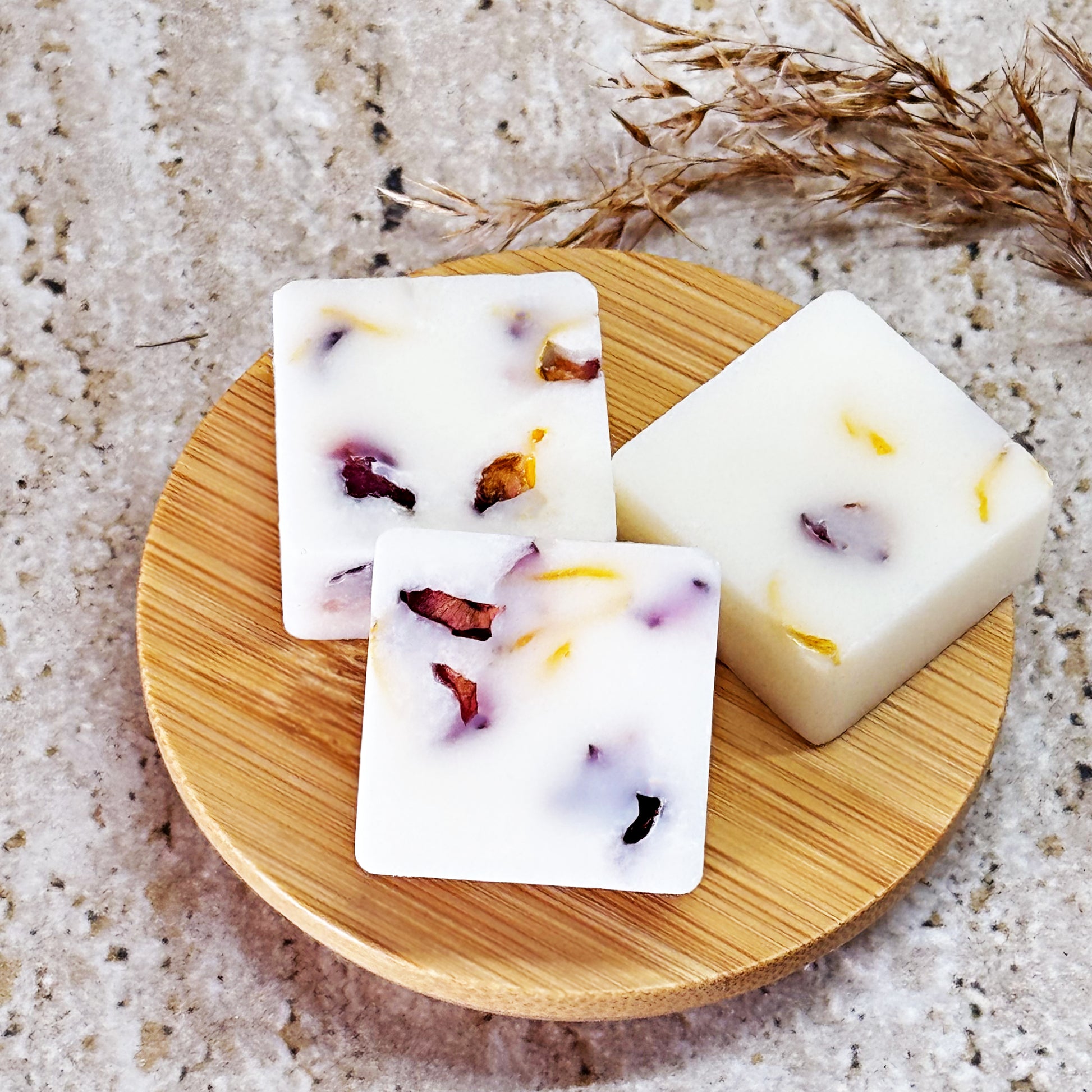 Three square botanical caramel vanilla latte wax melts with visible dried flower petals, displayed on a round wooden dish on a stone surface.