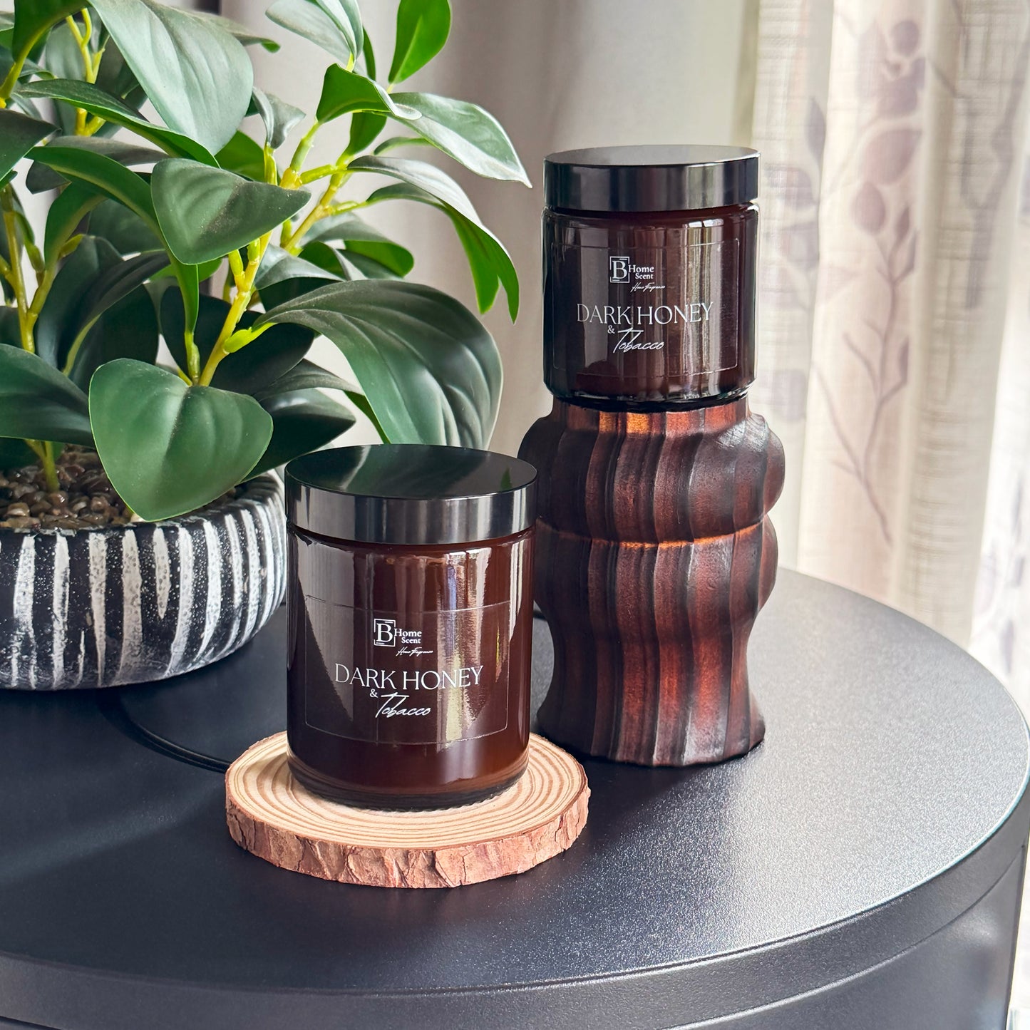 Two 'Dark Honey and Tobacco ' candles on a table with a plant in the background