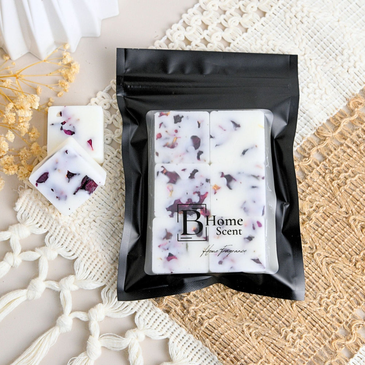A black resealable pouch containing three six rectangular wax melt bars with dried rose petals flower, shown on a styled surface with lace and woven mats. Two wax melts cubes are placed beside the pouch.
