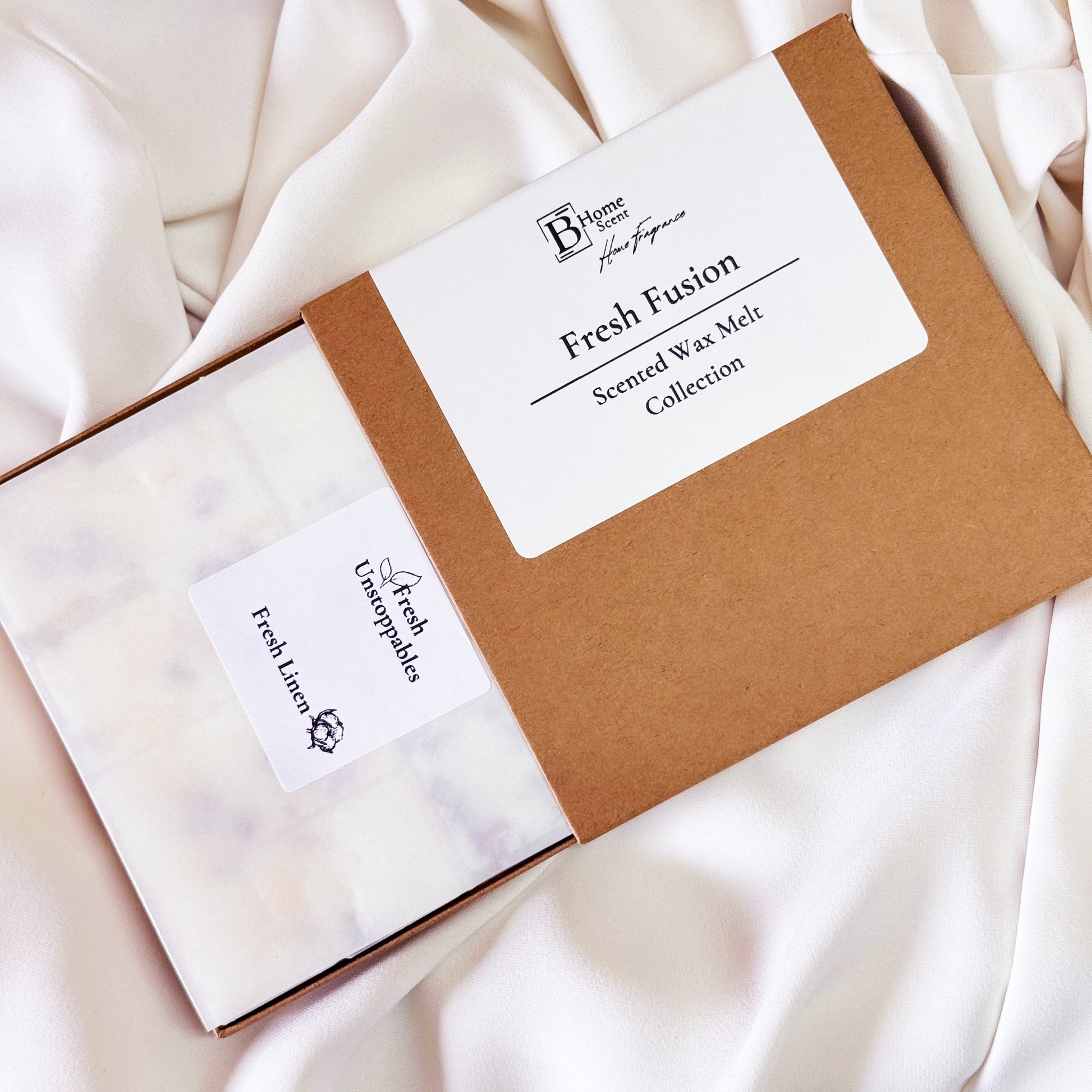 shows the Fresh Fusion Scented Wax Melt Collection from BHomeScent, placed on a soft, white fabric background. The brown box is partially open, revealing two sets of wax melts inside, each labelled individually. One label reads "Fresh Linen" and the other "Fresh Unstoppables." The packaging design is simple and clean, reflecting the fresh, laundry-inspired scents of the wax melts.