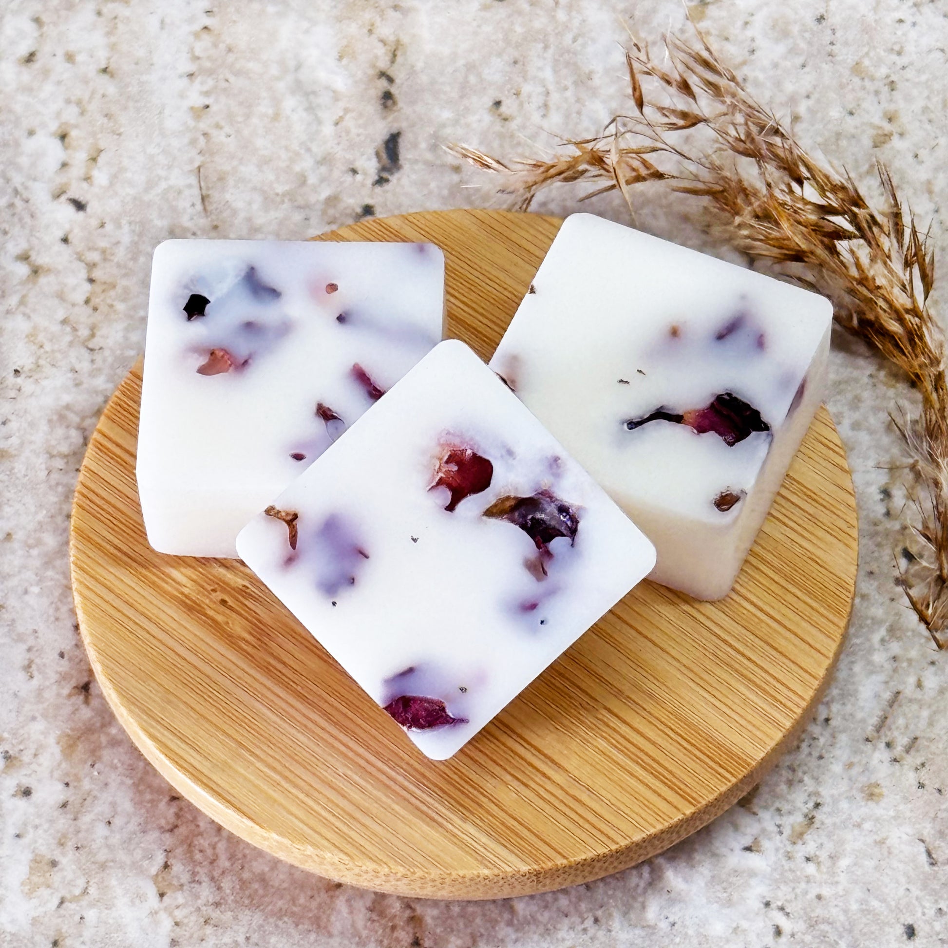 Hand holding a Cherry Blossom wax melt with dried rose petals, placing it into a white wax warmer. The packaged wax melts in a black resealable bag with the 'B Home Scent' logo are visible on a beige marble surface, alongside some rose petals and a small green leaf