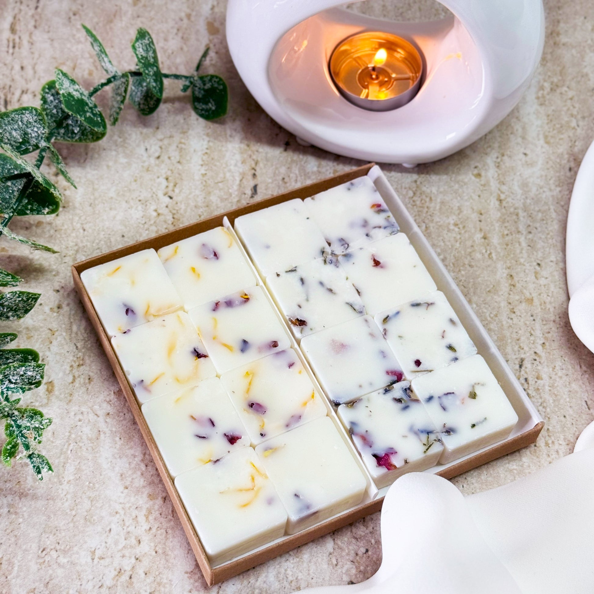 Fresh Fusion Scented Wax Melt Collection from BHomeScent, arranged on a light-coloured, textured surface. The box of wax melts is open, revealing two rows of neatly arranged wax cubes, each embedded with dried Lavender, Calendula and Rose Petals. To the right, a lit white wax melt burner adds a warm, cosy ambiance to the scene. A sprig of green leaves lies to the left, complementing the fresh, natural theme. The overall presentation emphasises the clean, laundry-inspired scents of the wax melts.