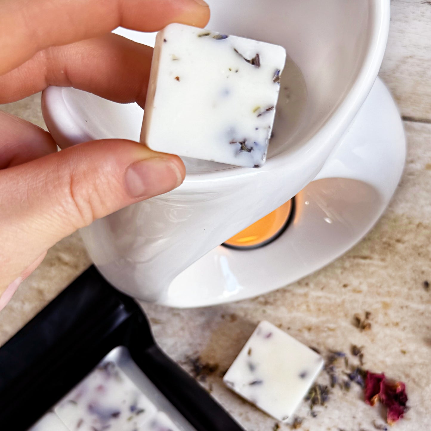 A hand placing a square botanical wax melt with embedded dried floral pieces into a white wax warmer