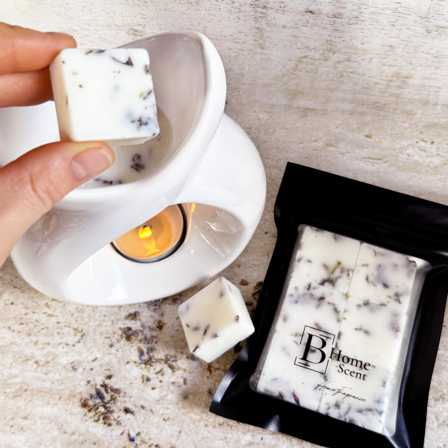 Hand holding a frosted eucalyptus wax melt with dried lavender, placing it into a white wax warmer. The packaged wax melts in a black resealable bag with the 'B Home Scent' logo are visible on a beige marble surface, alongside some lavender and a small green leaf