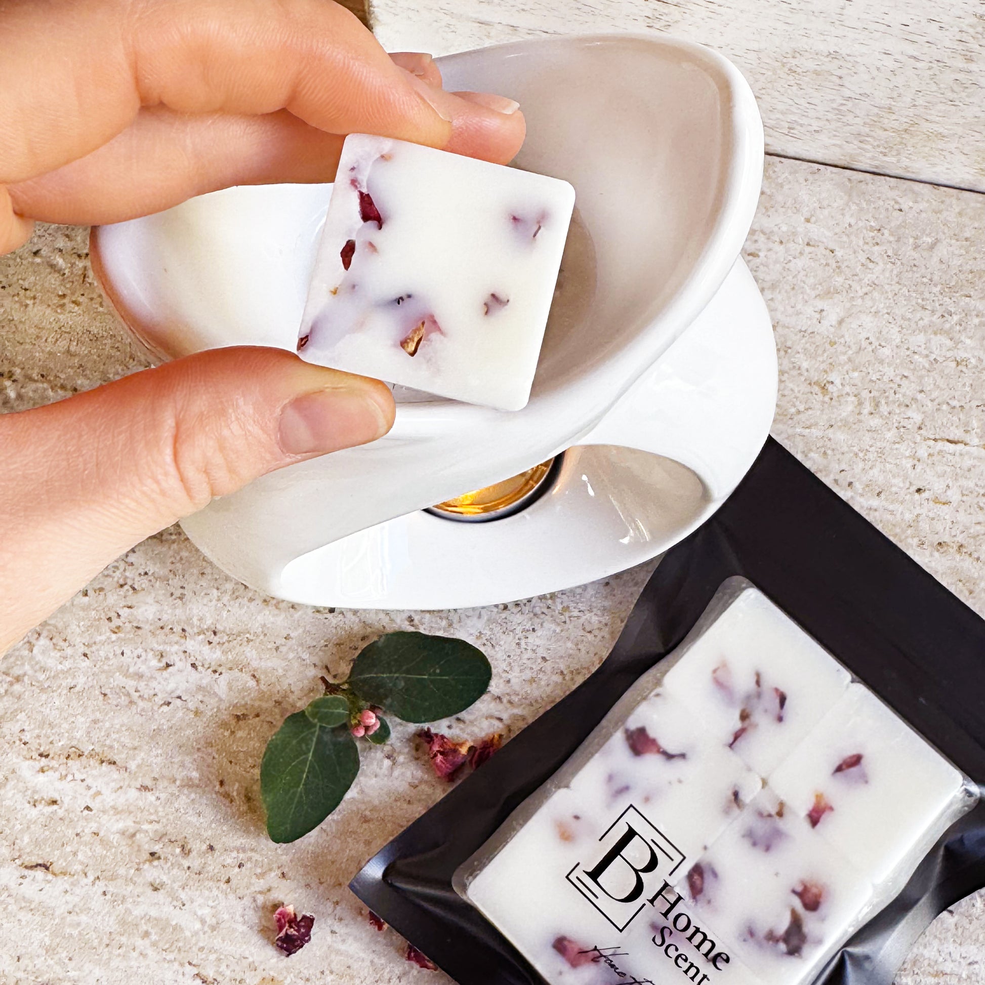 Hand holding a Cherry Blossom wax melt with dried rose petals, placing it into a white wax warmer. The packaged wax melts in a black resealable bag with the 'B Home Scent' logo are visible on a beige marble surface, alongside some rose petals and a small green leaf