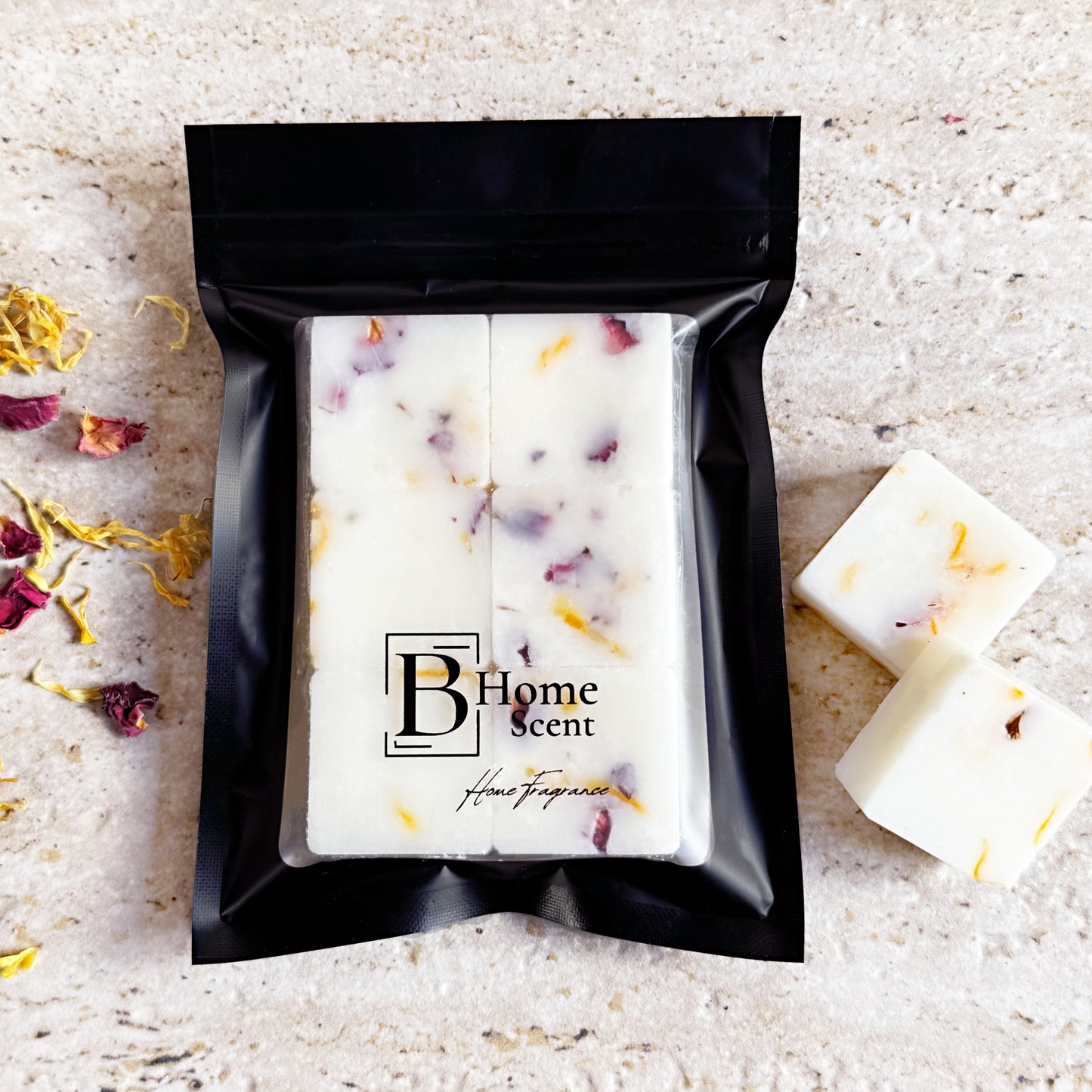 A black pouch labelled 'B Home Scent' containing Tobacco & Vanilla botanical wax melts with visible dried rose and calendula petals. Loose petals are scattered on the surface next to two wax melts