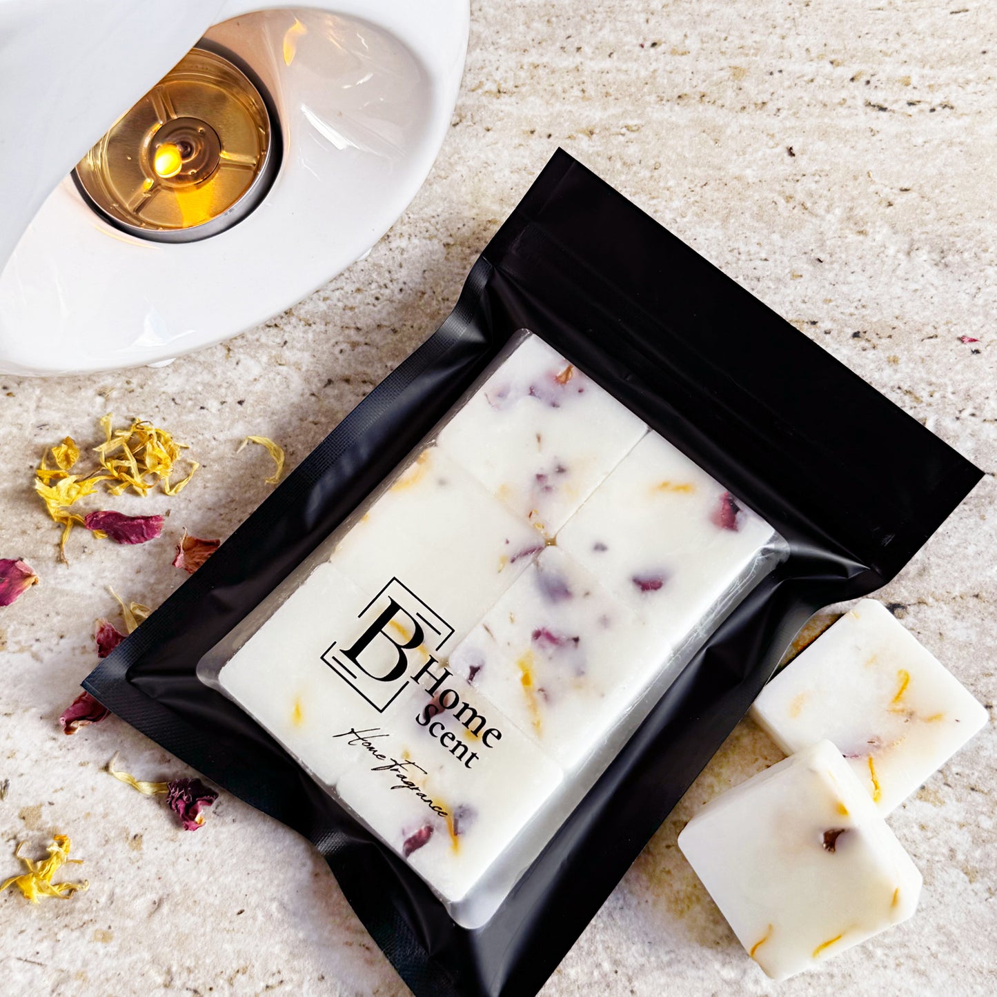 A black pouch of 'B Home Scent' Tobacco & Vanilla botanical wax melts, placed next to dried petals and a lit wax warmer, with loose melts visible on the side.