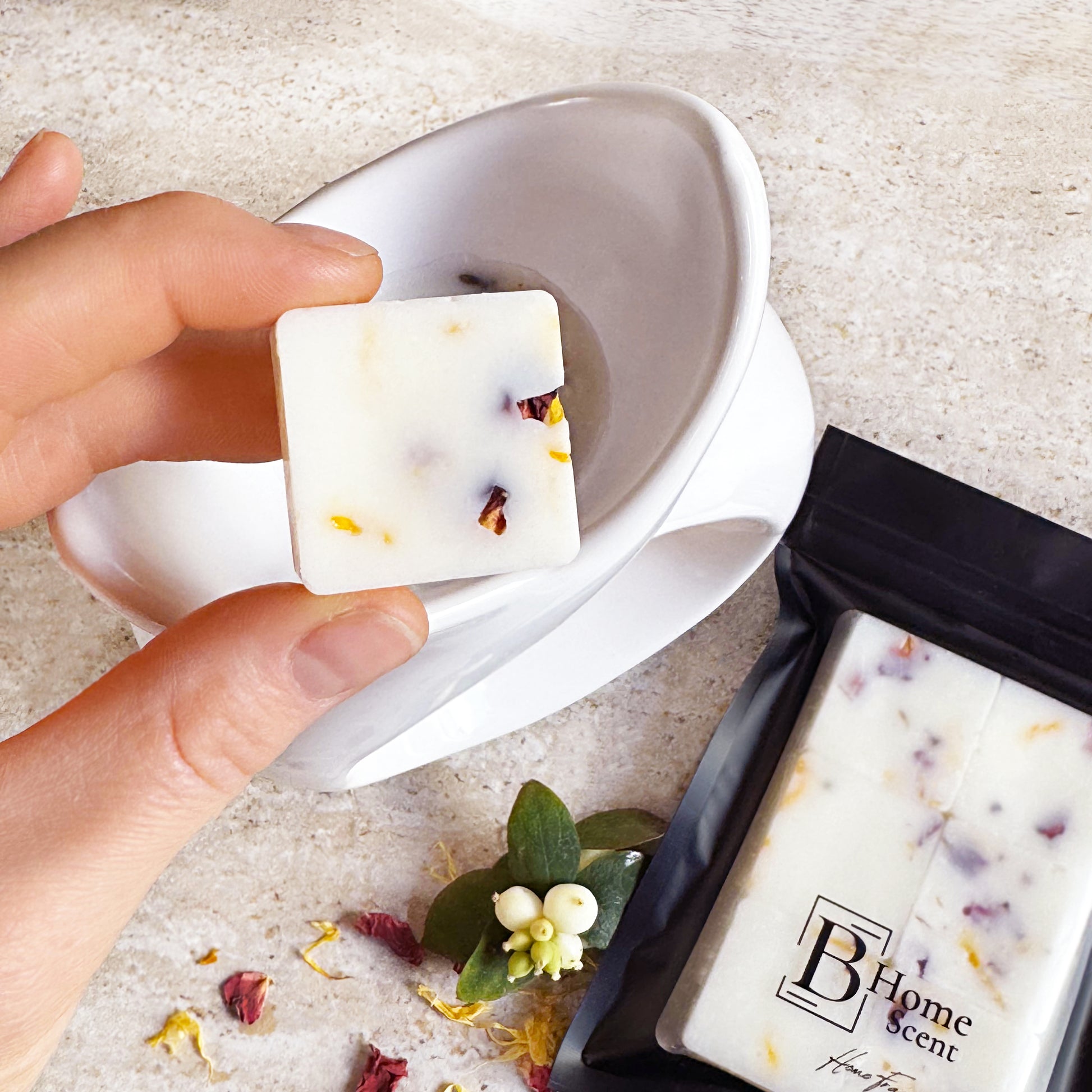 A hand holding a Tobacco & Vanilla wax melt with dried rose and calendula petals, placing it into a white wax warmer. A black pouch of wax melts is visible in the background.