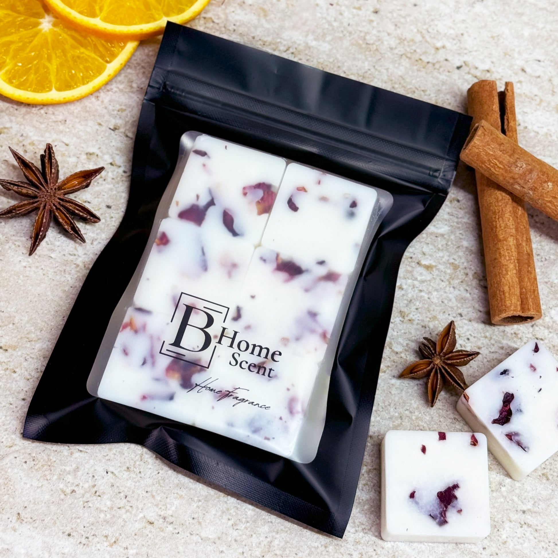 A black pouch of 'B Home Scent' Christmas Spice botanical wax melts with dried rose petals, displayed on a marble surface surrounded by cinnamon sticks, star anise, and orange slices.