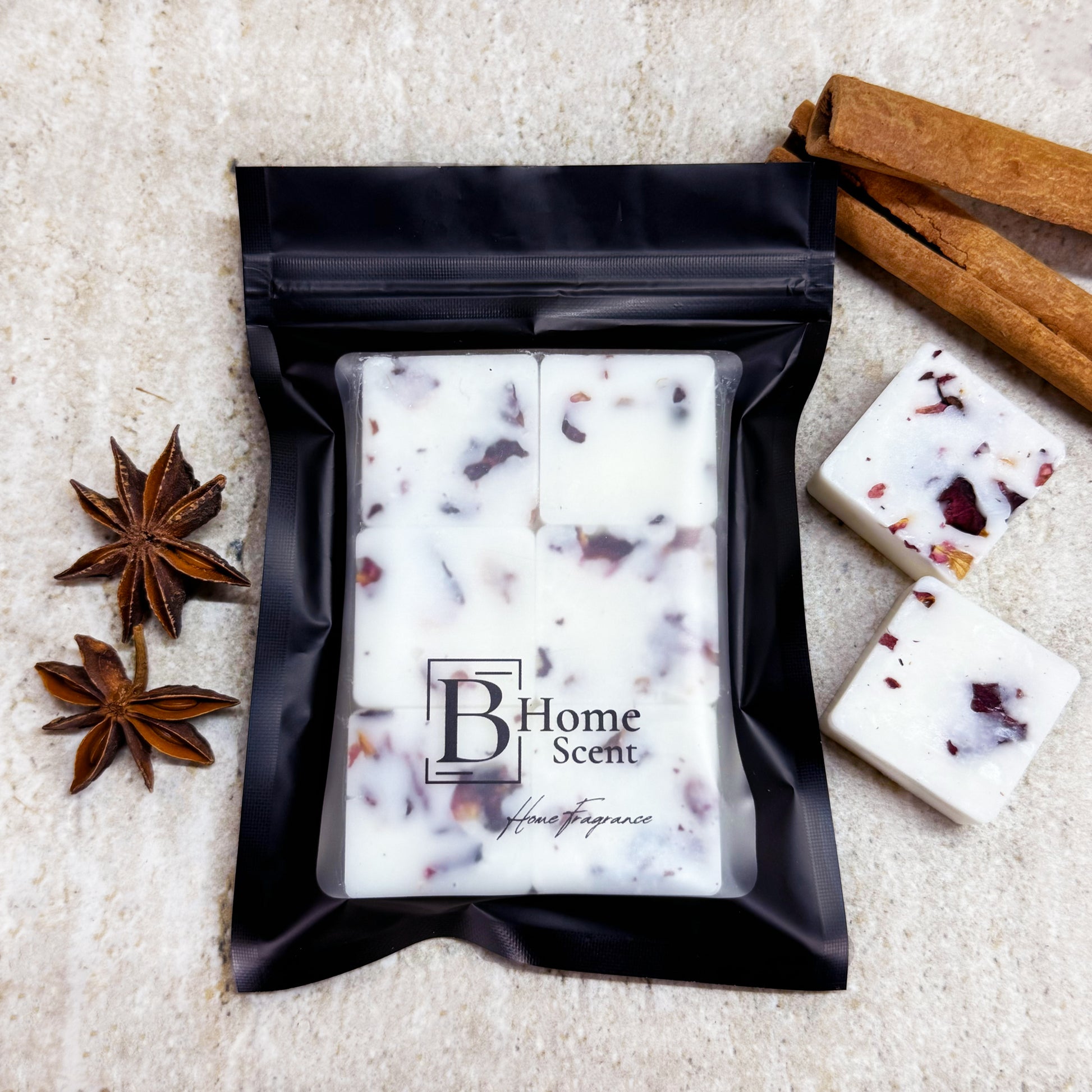 A black pouch labelled 'B Home Scent' containing six Christmas Spice botanical wax melts with dried rose petals, placed on a marble surface next to cinnamon sticks, star anise, and loose wax melts.