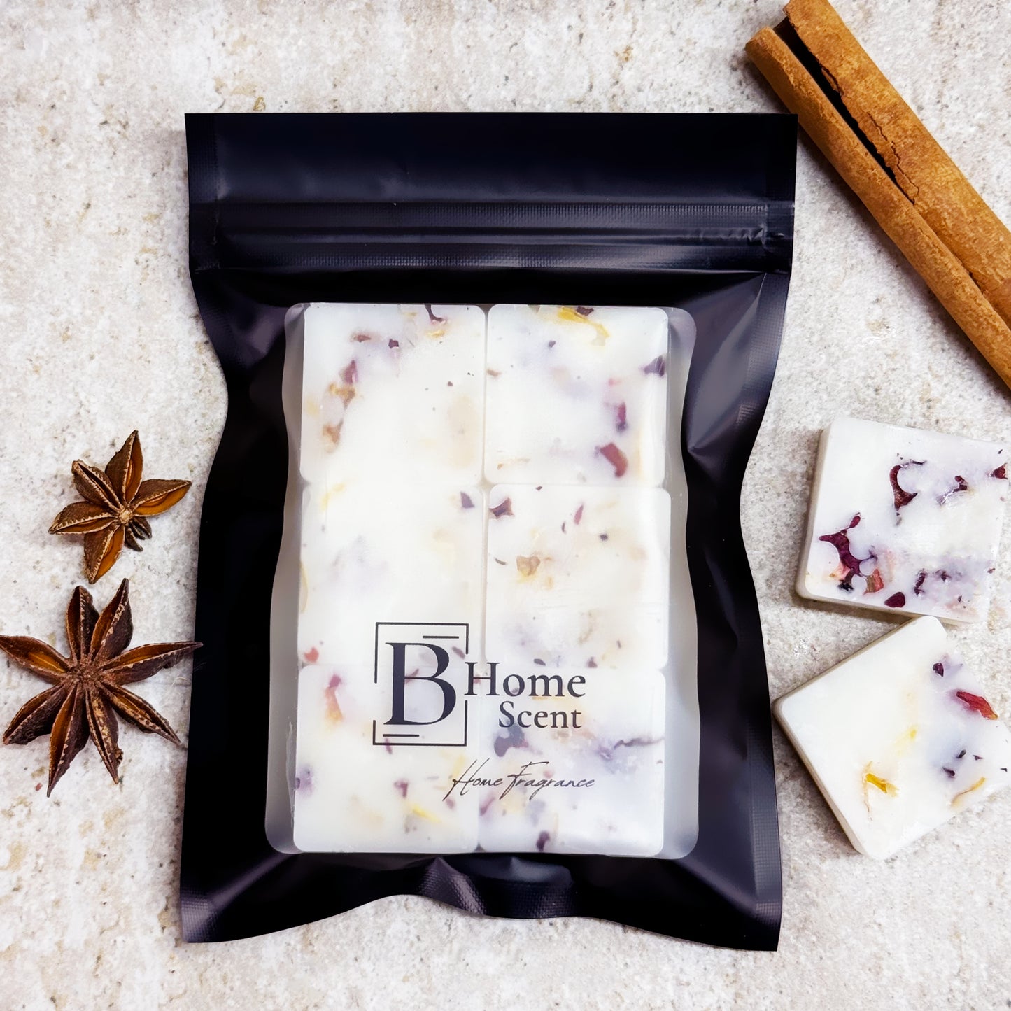 A black pouch of 'B Home Scent' Gingerbread botanical wax melts with dried rose and calendula petals, placed on a marble surface alongside cinnamon sticks and star anise.