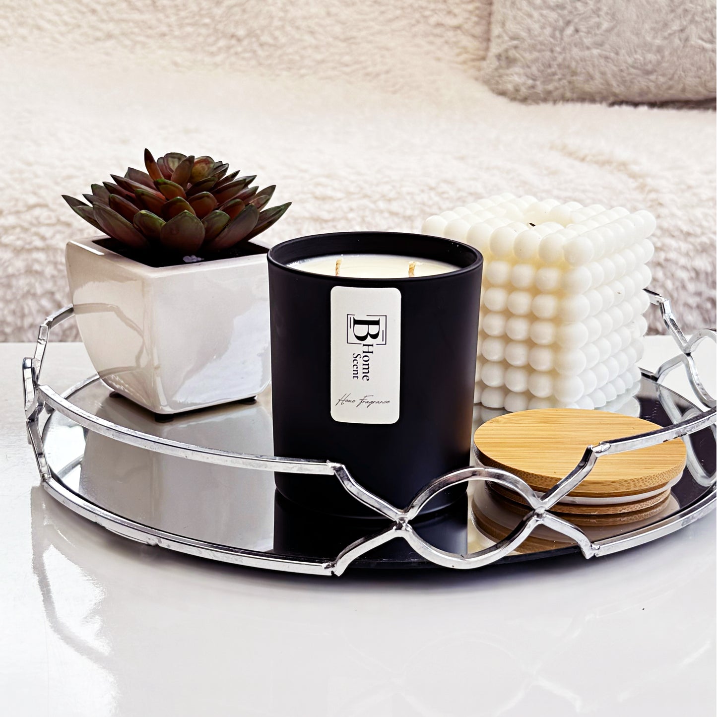 A black Coffee Cake scented candle with two wicks, labelled 'B Home Scent,' placed on a mirrored tray alongside a small potted succulent and a decorative bubble candle. A wooden lid rests next to the candle, with a cosy blanket in the background.