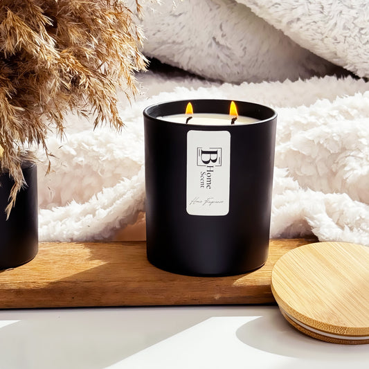 A lit Coffee Cake scented candle with two wicks in a black jar, labelled 'B Home Scent,' placed on a wooden tray, with a cosy blanket in the background and a wooden lid beside the candle.