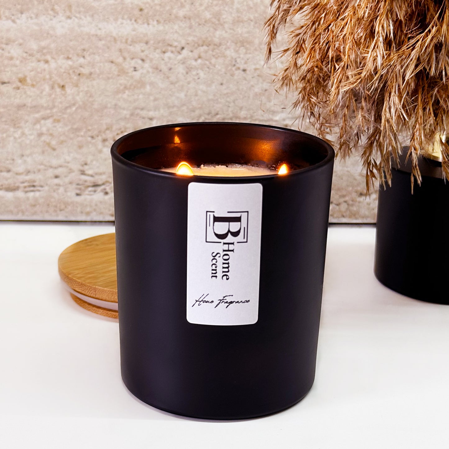 "Black matte candle jar with a wooden lid, featuring a double wick for an even burn. The label reads 'B Home Scent - Home Fragrance'. The candle is placed on a white surface with dried pampas grass decor in the background, creating a warm and cosy ambiance."