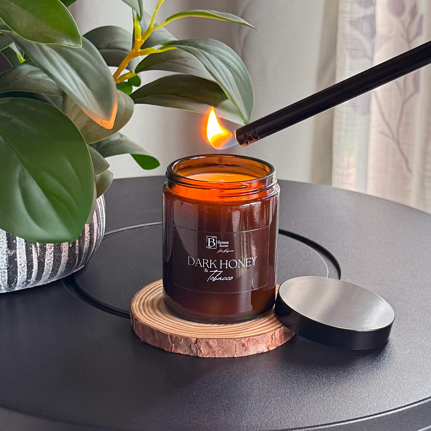 Candle labelled 'Dark Honey and Tobacco' being lit on a wooden coaster with a plant in the background.