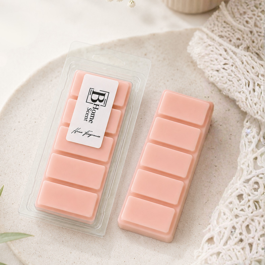 A package of six pale pink Myrrh and Tonka wax melt snap bar next to an unpackaged  wax melt snap bar, both labelled 'B Hone Scents', displayed on a textured beige stone plate with a white lace cloth.