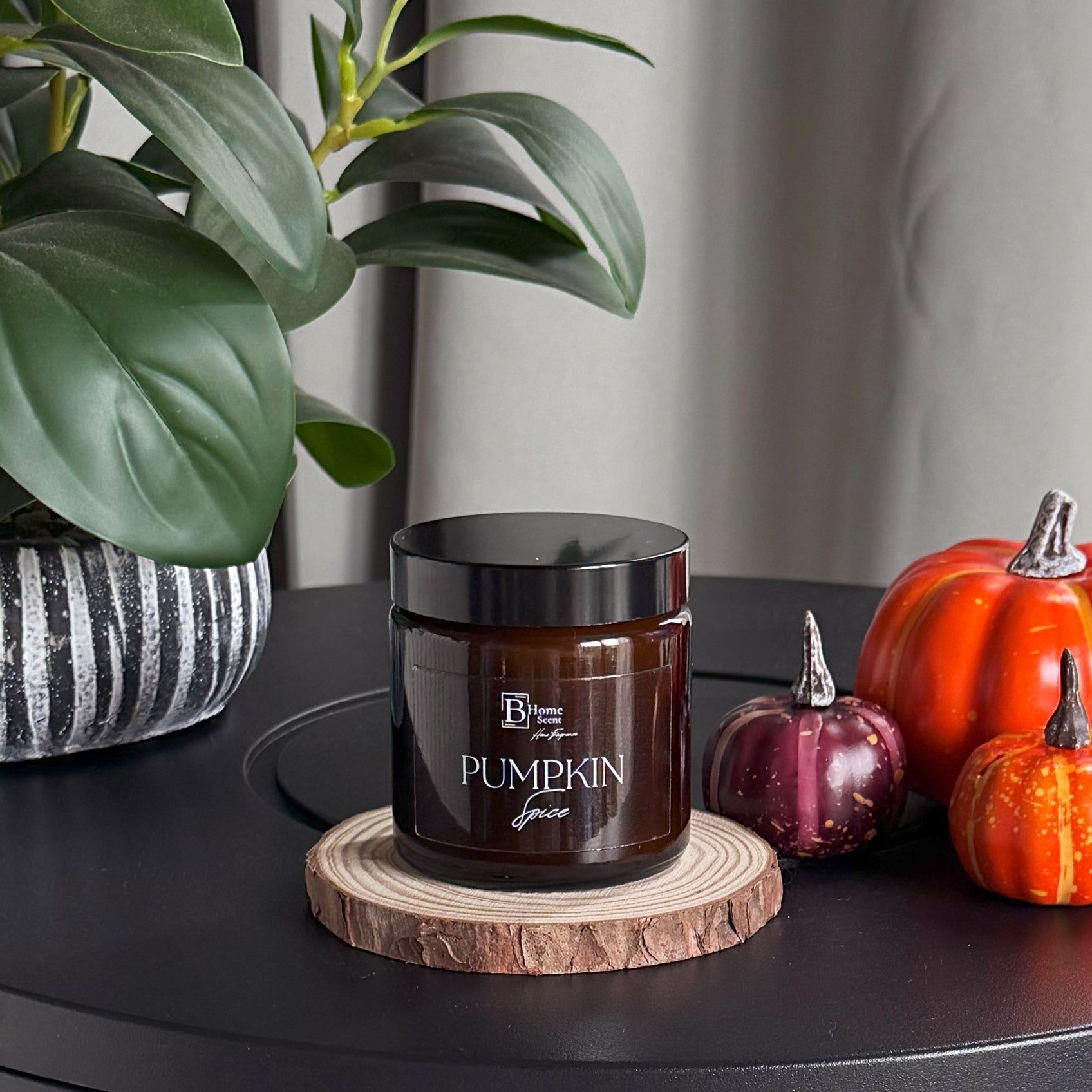 Jar of pumpkin spice on a wooden coaster with decorative pumpkins and a plant in the background.