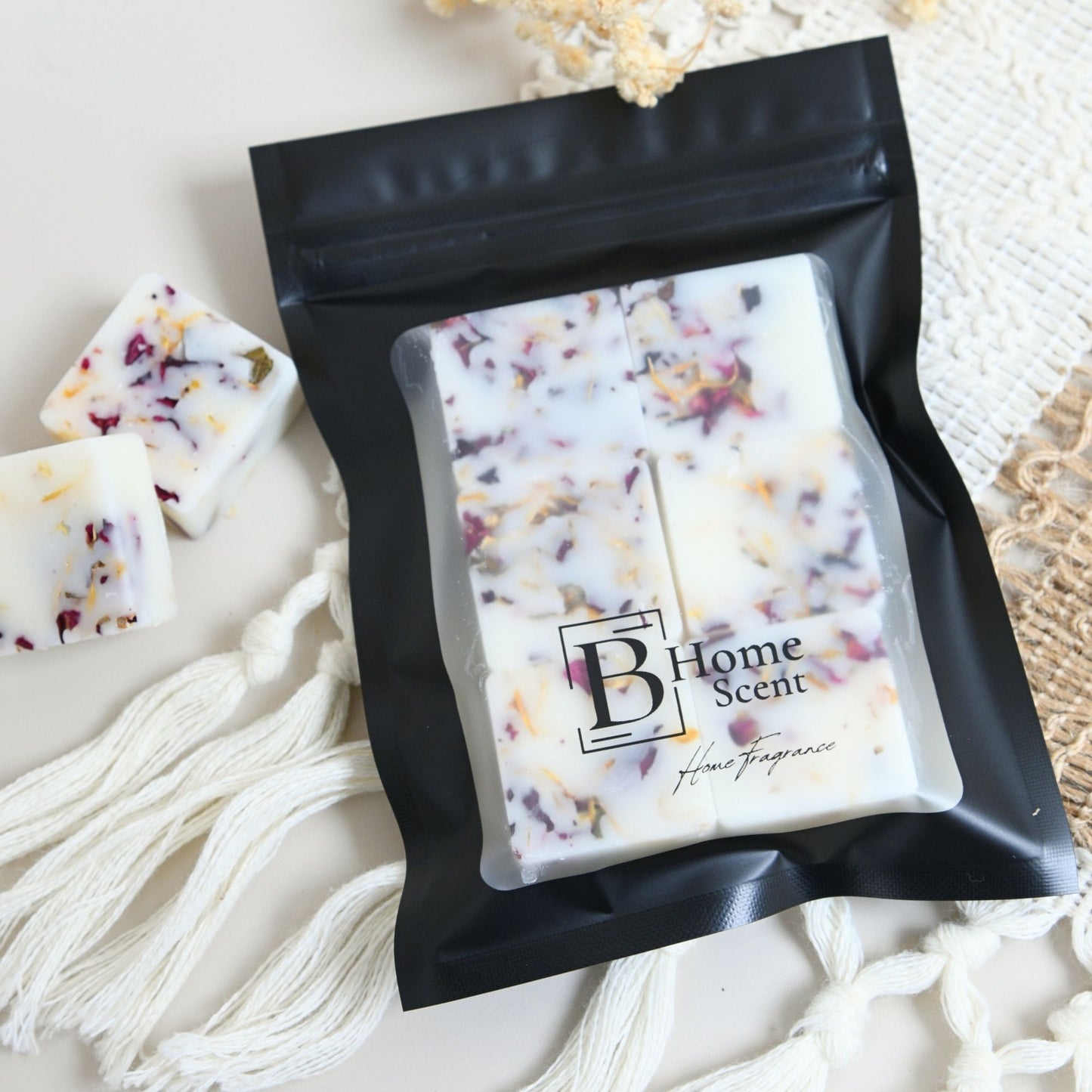 A black resealable pouch containing six square home fragrance wax melts, with a few loose white wax pieces beside it. The pouch has a clear front window and a printed logo reading 'B Home Scent'.