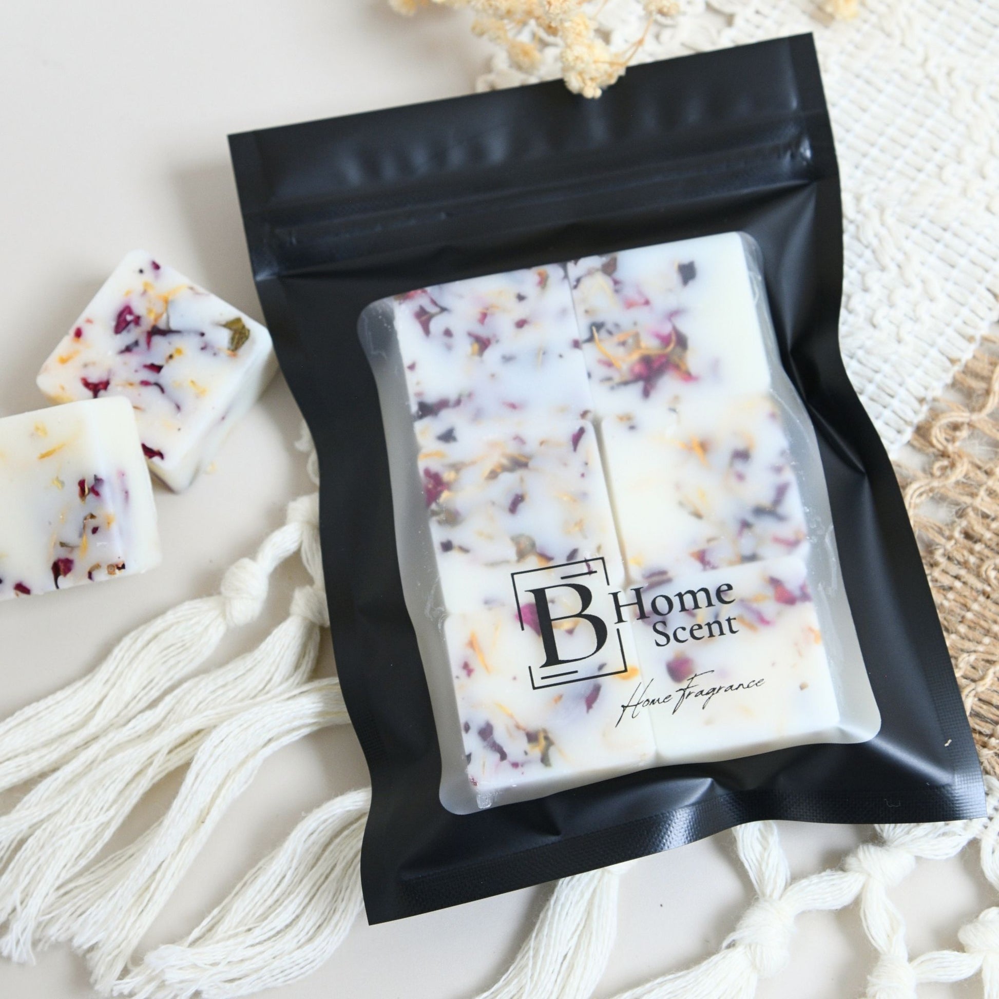 A black resealable pouch containing six square home fragrance wax melts, with a few loose white wax pieces beside it. The pouch has a clear front window and a printed logo reading 'B Home Scent'.