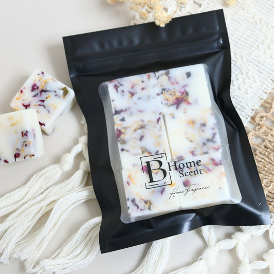 A black resealable pouch containing six square home fragrance wax melts, with a few loose white wax pieces beside it. The pouch has a clear front window and a printed logo reading 'B Home Scent'.