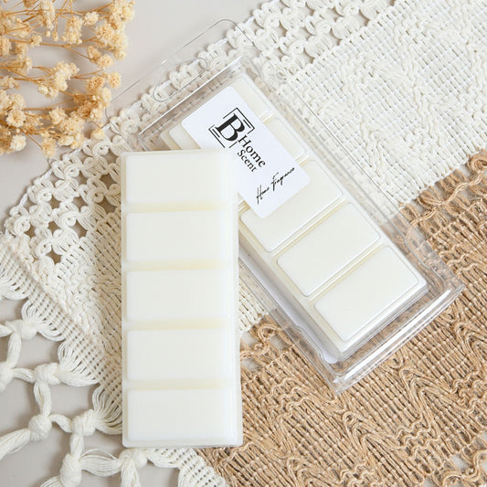 A set of white wax melts, with one unwrapped snap bar and one packaged in a clear plastic clamshell container, styled on a beige and white boho lace background with dried flowers.