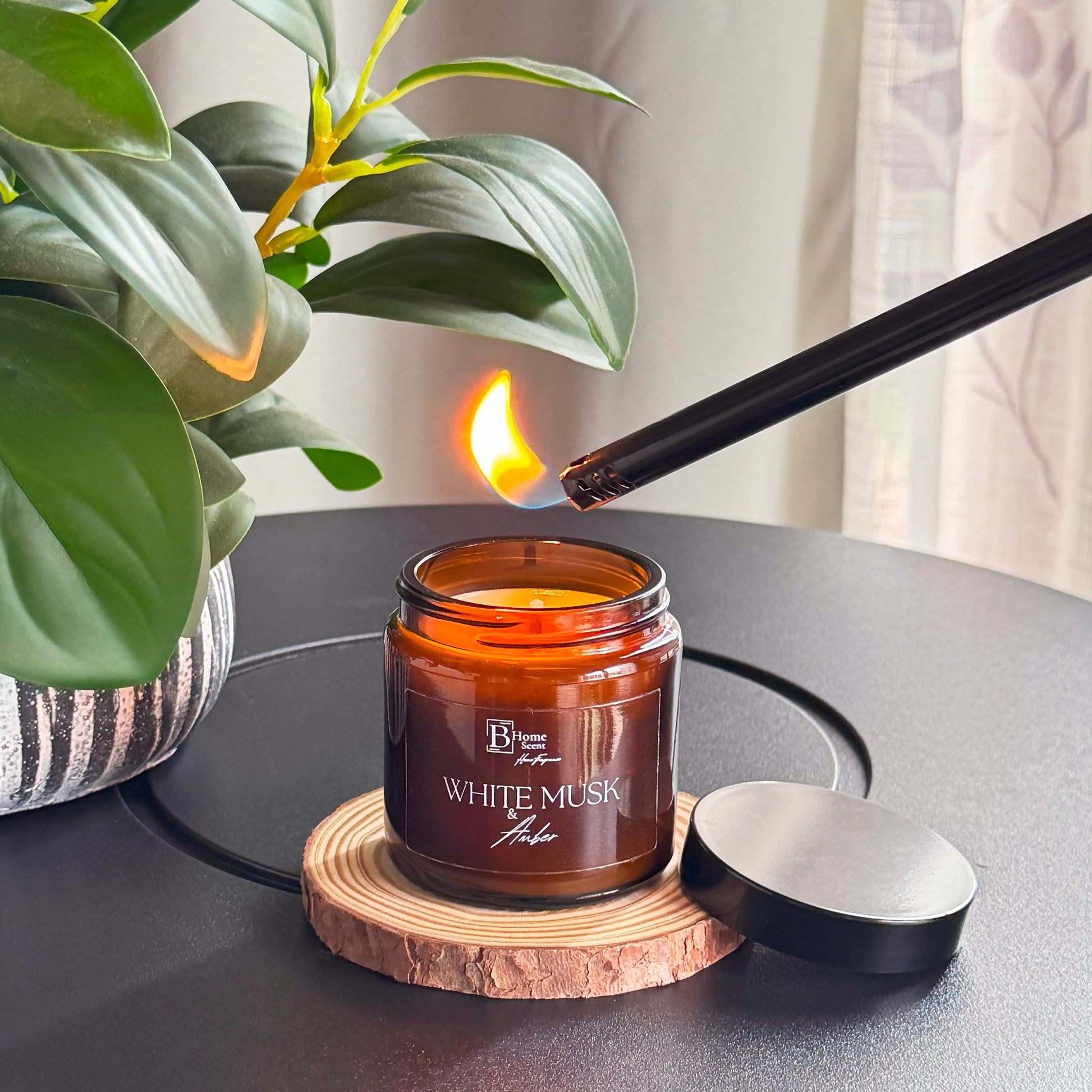 Candle labeled 'White Musk' being lit with a match on a dark surface next to a plant.