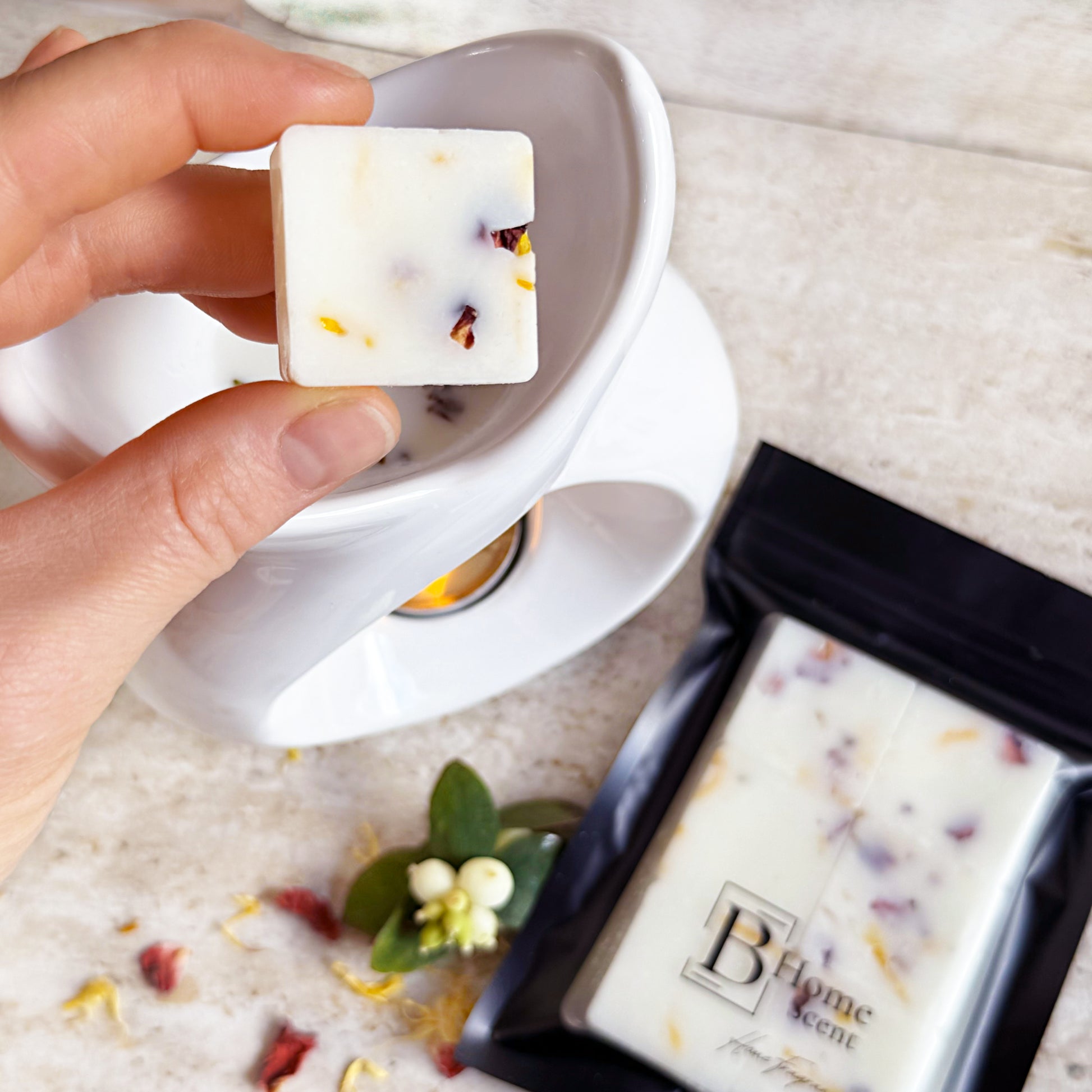 Amber and Sweet Orange Botanical Wax Melts -Hand holding a square wax melt with dried flowers, surrounded by more wax melts and decorative elements on a textured surface.