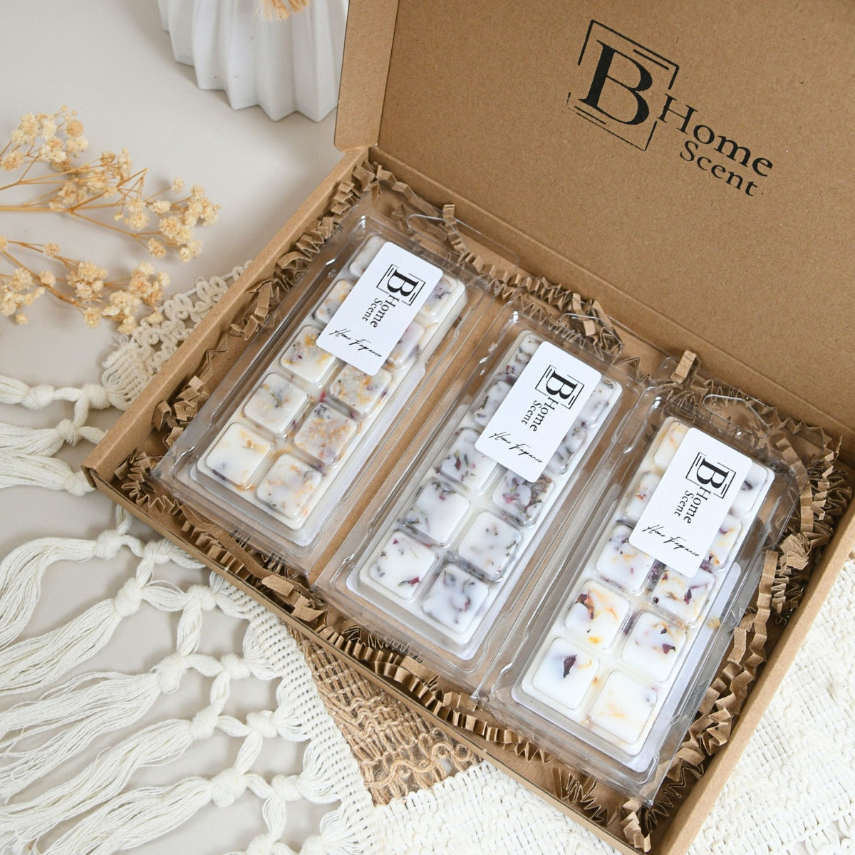 An open kraft gift box of Autumn Wax Melt Snap Bar Collection, containing three clear plastic packages of wax melts filled with white wax and small botanical inclusions, arranged in brown paper shred with dried flowers and a white knit textile around it.