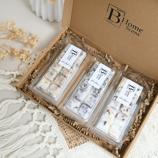 An open kraft gift box of Autumn Wax Melt Snap Bar Collection, containing three clear plastic packages of wax melts filled with white wax and small botanical inclusions, arranged in brown paper shred with dried flowers and a white knit textile around it.
