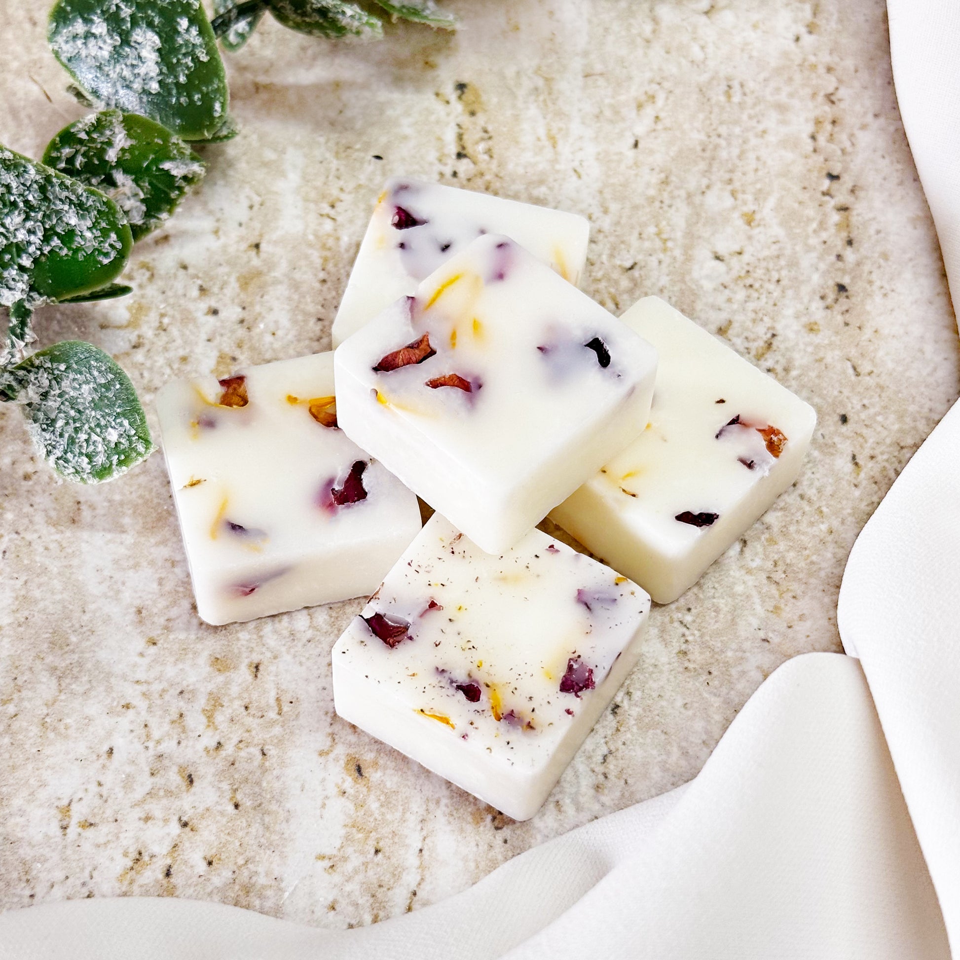 Five square gingerbread botanical wax melts with dried rose and calendula petals, arranged on a marble surface with frosted green leaves in the background, and a white fabric draped on the side.