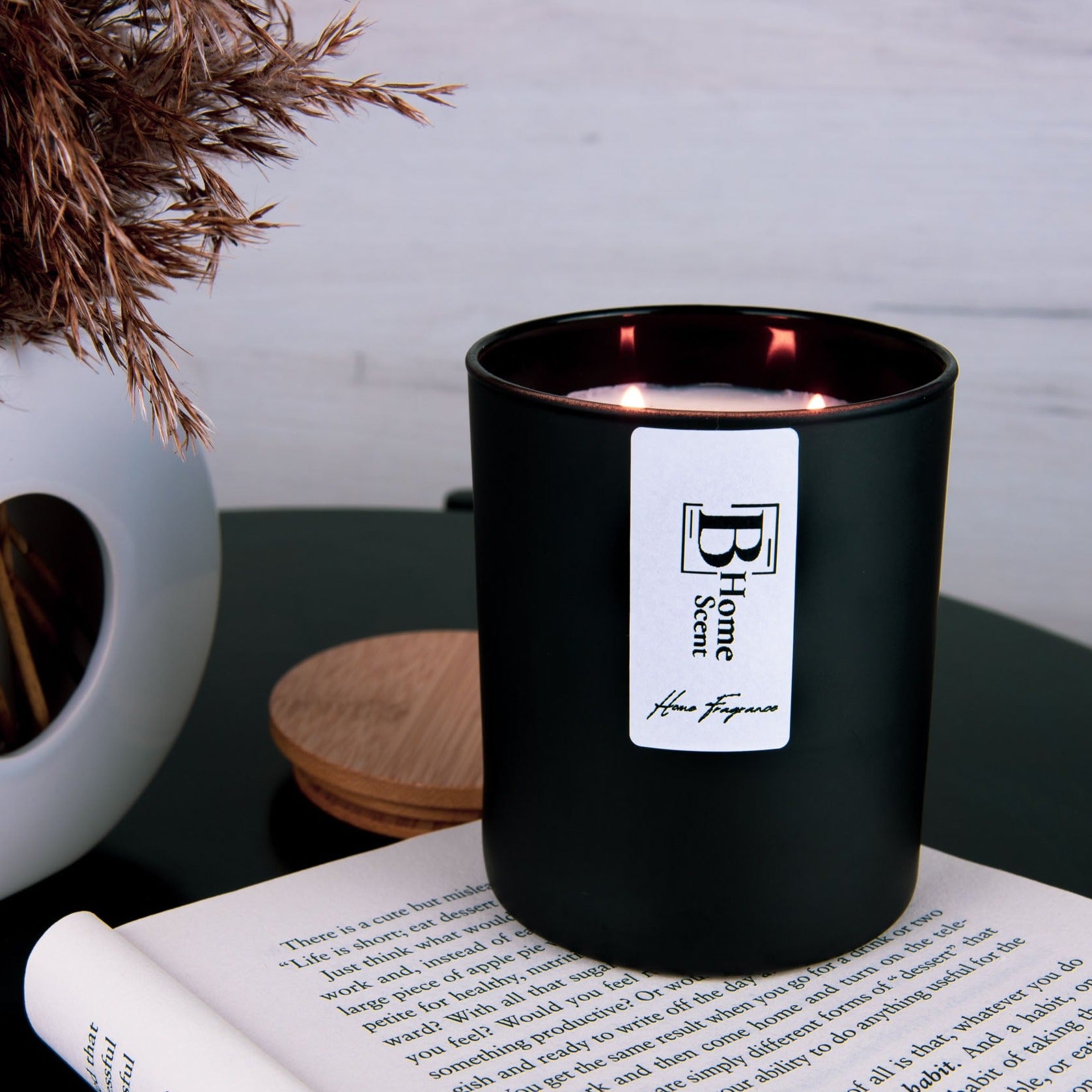 Black matte candle jar with a white label reading 'B Home Scent - Home Fragrance, set on a dark green surface next to an open book. The candle is complemented by a modern white vase holding dried pampas grass, creating a cosy and inviting atmosphere, perfect for relaxation and reading.