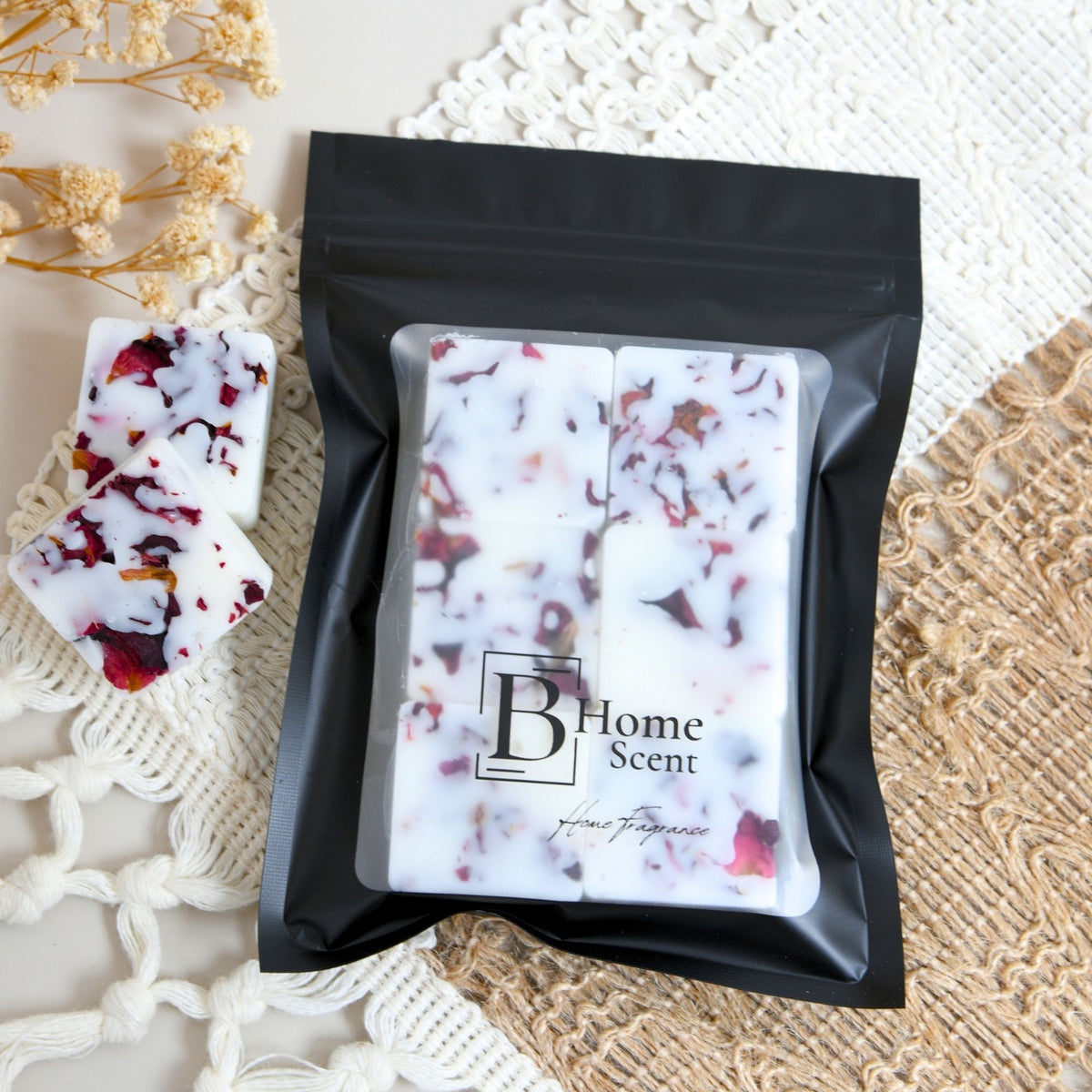 Myrrh and tonka wax melts in a black resealable pouch containing six white square home scent cubes with red dried flower pieces, placed on a cream lace surface with a woven mat and small dried flowers nearby. The pouch label reads 'B Home Scent'.