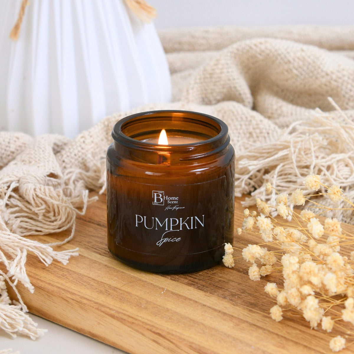 Pumpkin Spice Scented Candle in an amber jar with a lit wick, surrounded by dried flowers on a wooden surface.