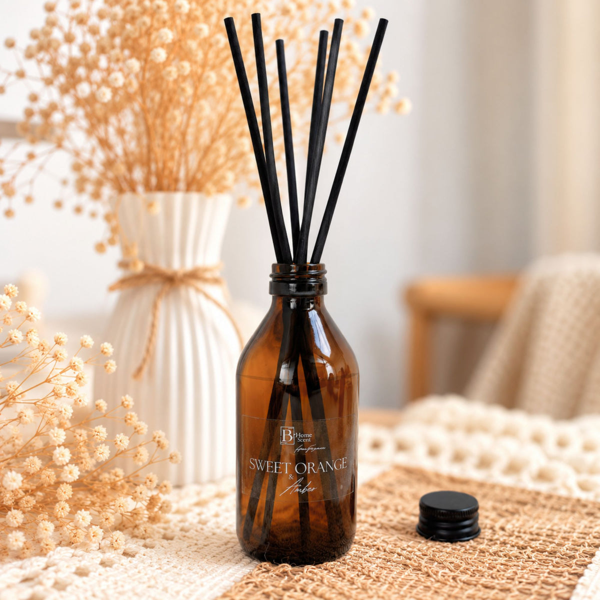 sweet orange and amber reed diffuser bottle with black reeds and a black cap, labelled Sweet Orange and amber, styled on a woven surface with dried flowers and a soft indoor background.
