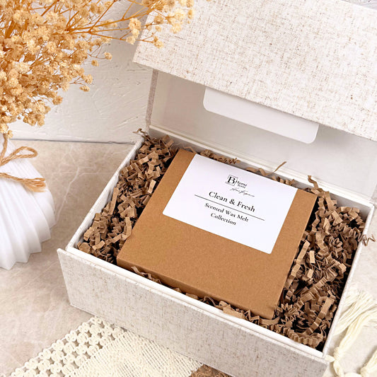 A square brown gift box labeled 'Clean & Fresh' a sits inside an open textured cream-colored box filled with brown crinkle paper shred. The scene is styled with dried flowers and a white candle on a light beige surface.